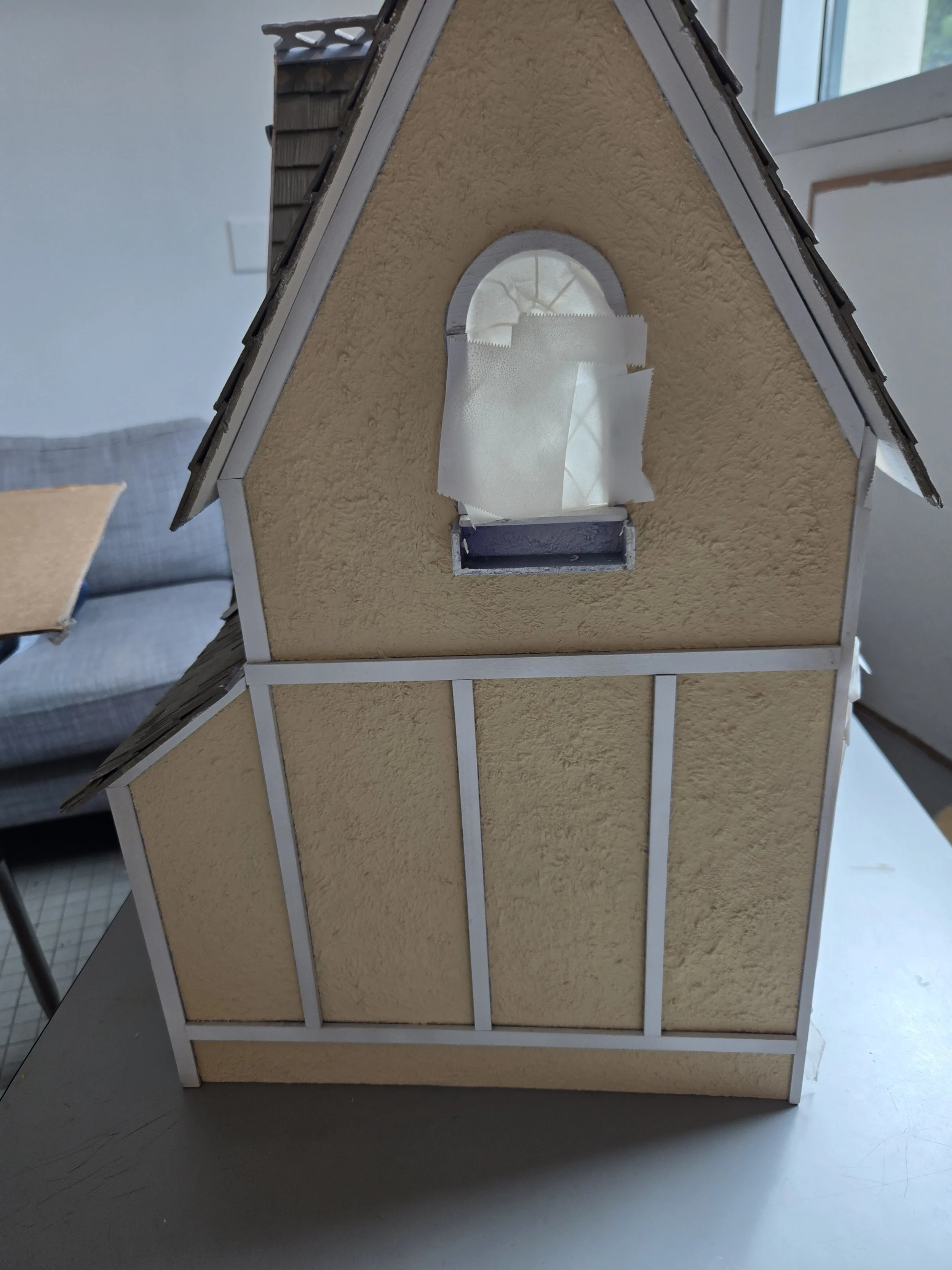 Model of a house with a sloped roof, beige textured walls, a small arched window covered with paper, and a brick-shaped door opening below the window.