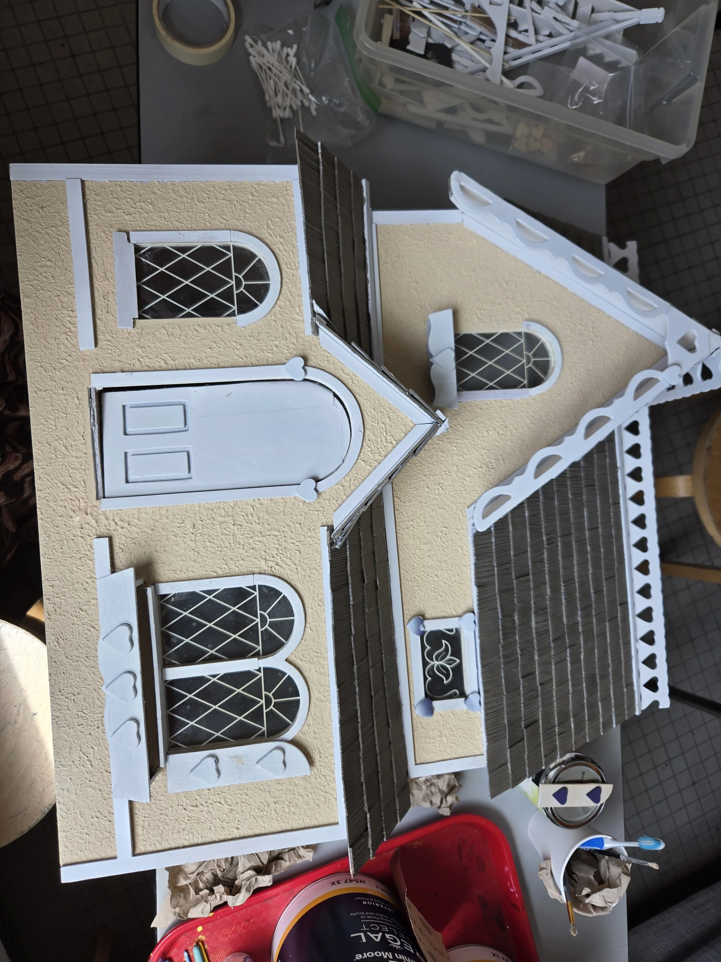 A work area with a miniature house model, tools, and art supplies. The house has beige textured walls, arched windows with decorative grilles, and a front door. There are various craft materials on the table, including a container of tiny hearts, bru