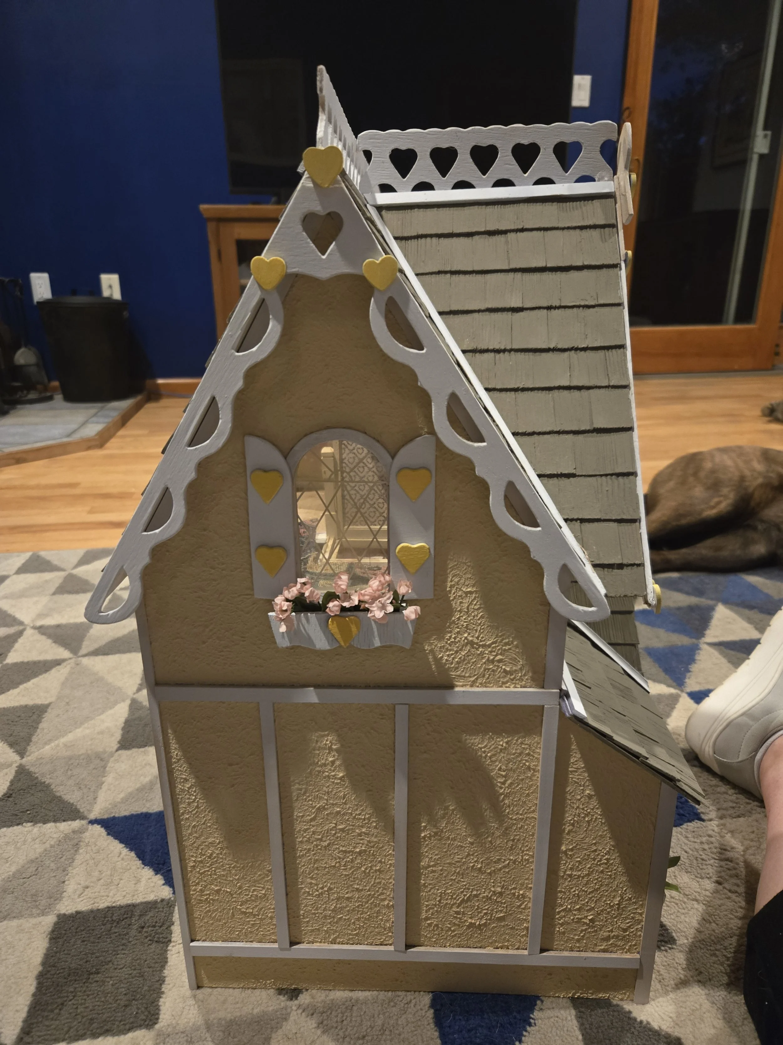 Miniature decorative house with heart-shaped cutouts and a window with pink flowers in a window box, placed indoors on a beige wall with a multicolored patterned rug in front.