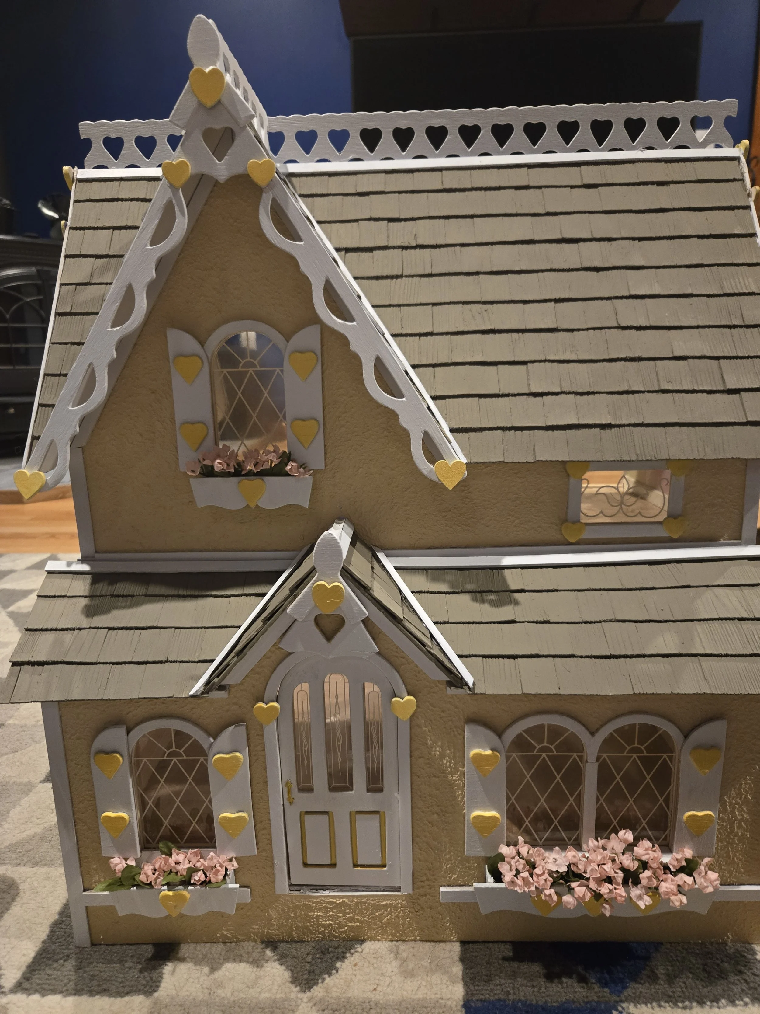 A dollhouse with cream-colored walls, white trim, heart-shaped decorations, pink flowers in window boxes, and a shingled roof.
