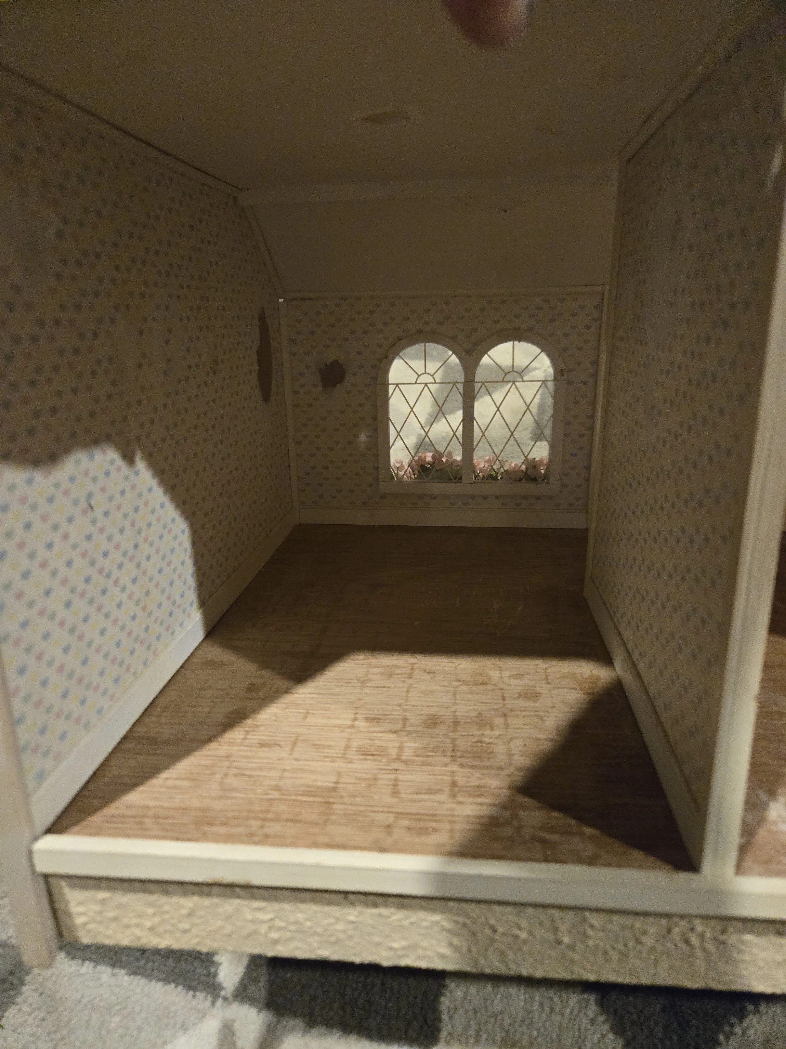 Interior view of a dollhouse with a window, floral curtains, and patterned wallpaper.