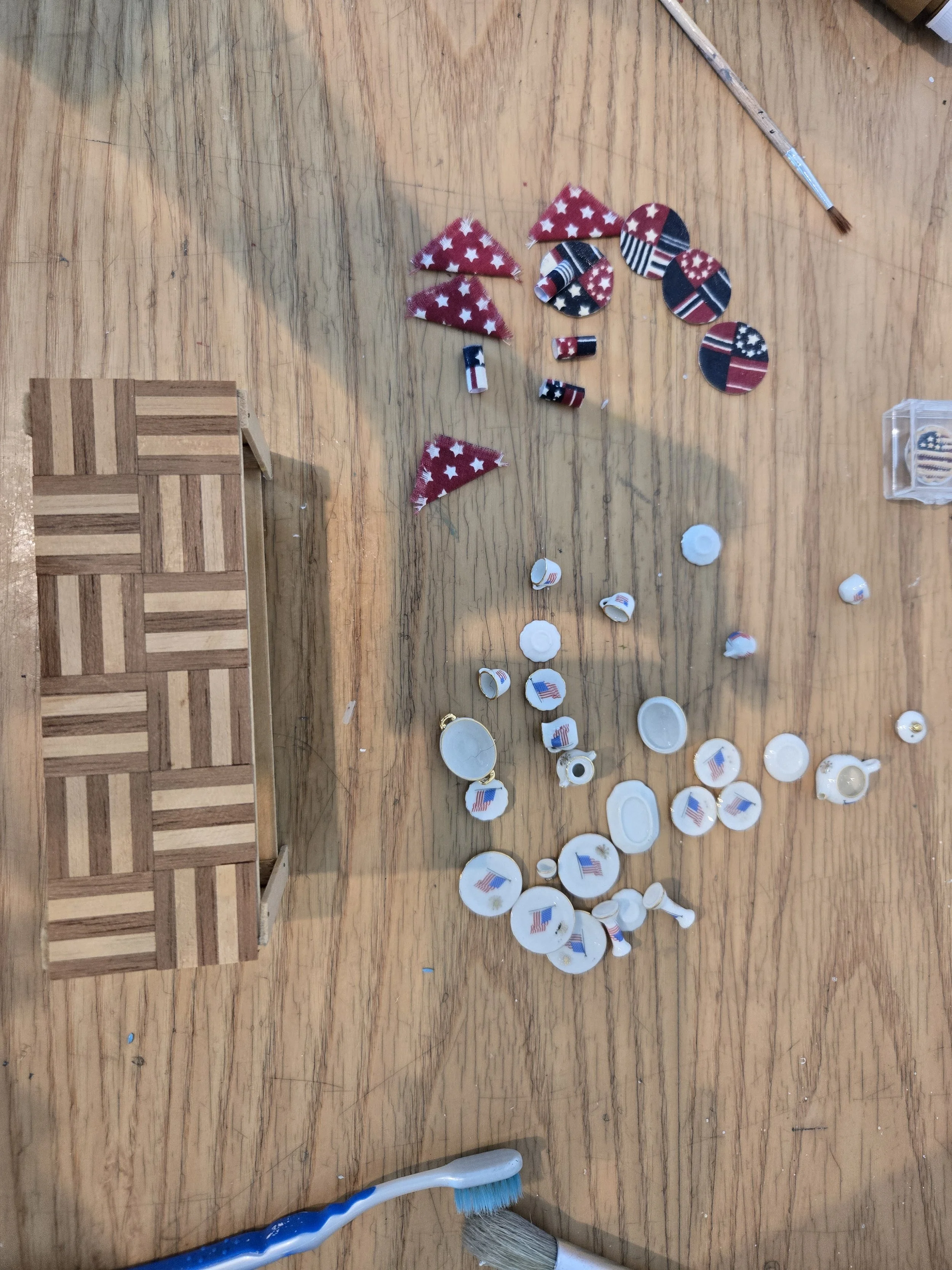 A wooden table with broken pieces of circular and triangular objects decorated in red, white, and blue patriotic patterns, possibly decorations or confetti. There is a small wooden box on the left and cleaning tools, including a toothbrush and a pain