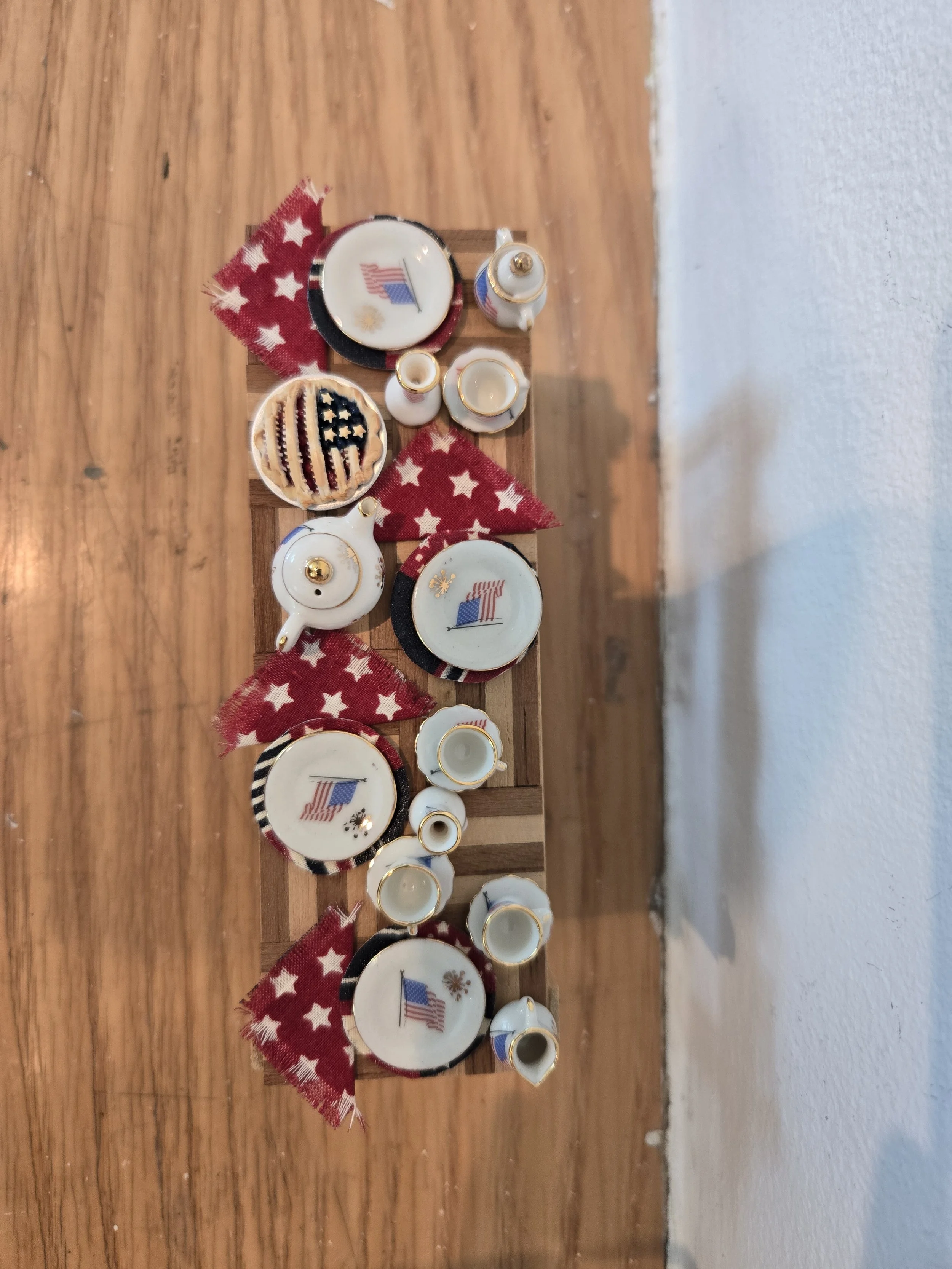 Miniature patriotic tea set with plates, cups, and teapot, decorated with American flag designs, displayed on a wooden surface with star-patterned fabric.
