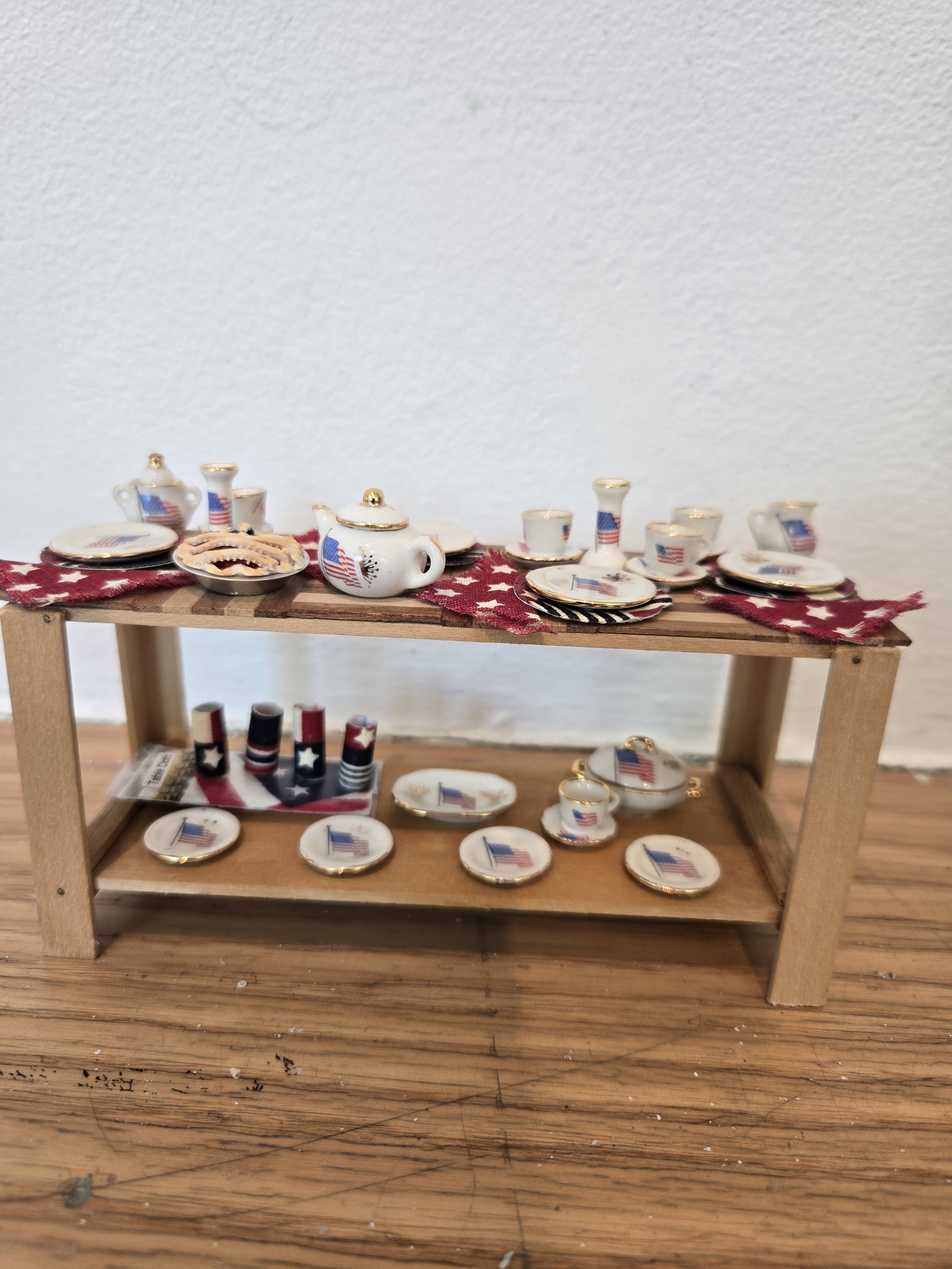 Miniature ceramic tea set with American flags on cups, saucers, teapot, and sugar bowl, arranged on a wooden shelf with a red, white, and blue patriotic table runner.