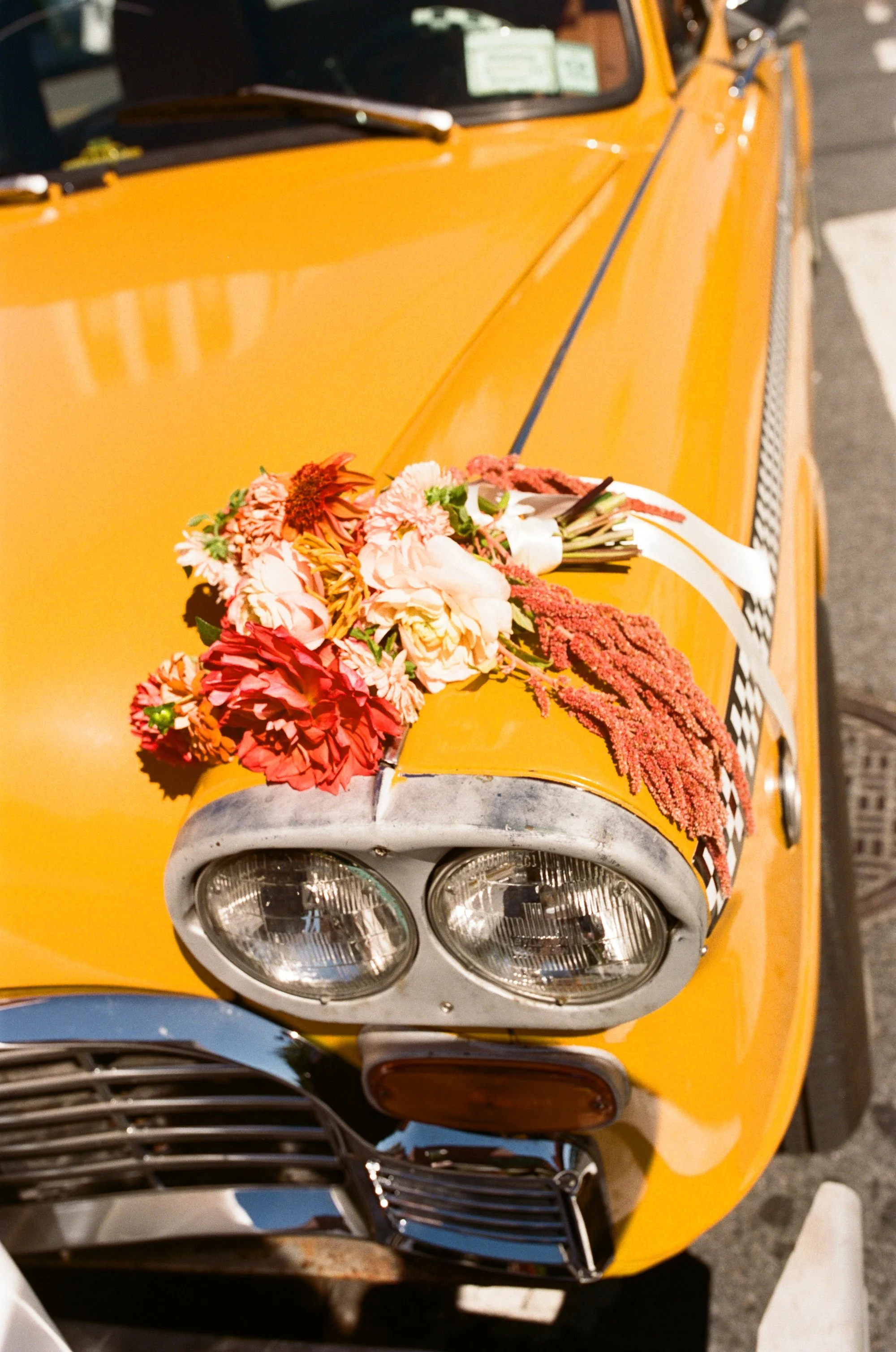 The front of a vintage yellow car decorated with a bouquet of pink, red, and cream-colored flowers on the hood.