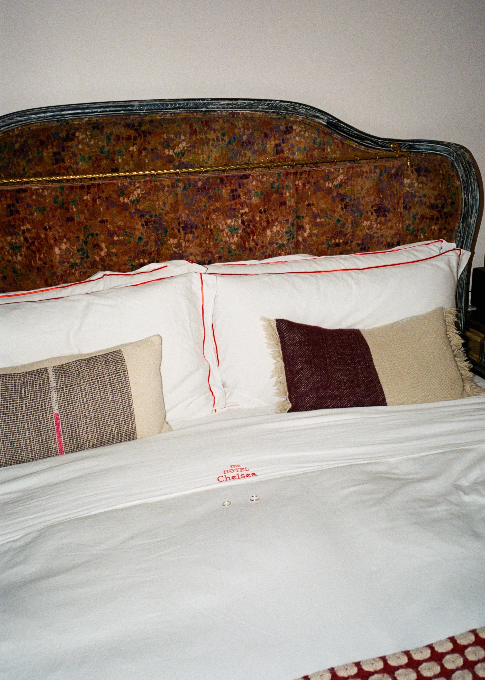 A bed with white sheets and pillows, two decorative pillows with different patterns, and an ornate headboard with a multicolored design, located in a hotel room.