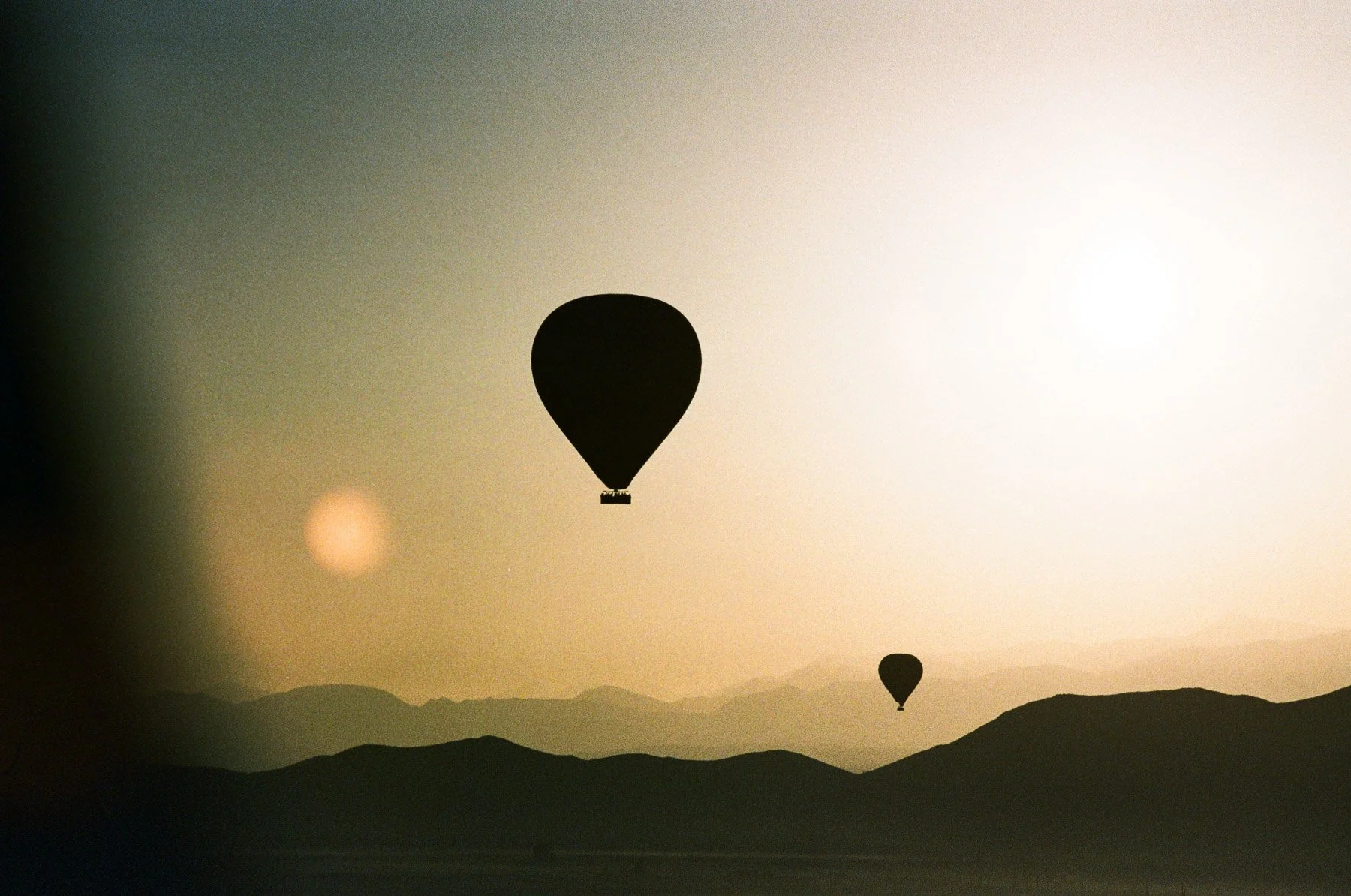 Silhouettes of hot air balloons drifting over a mountainous landscape at sunset or sunrise.