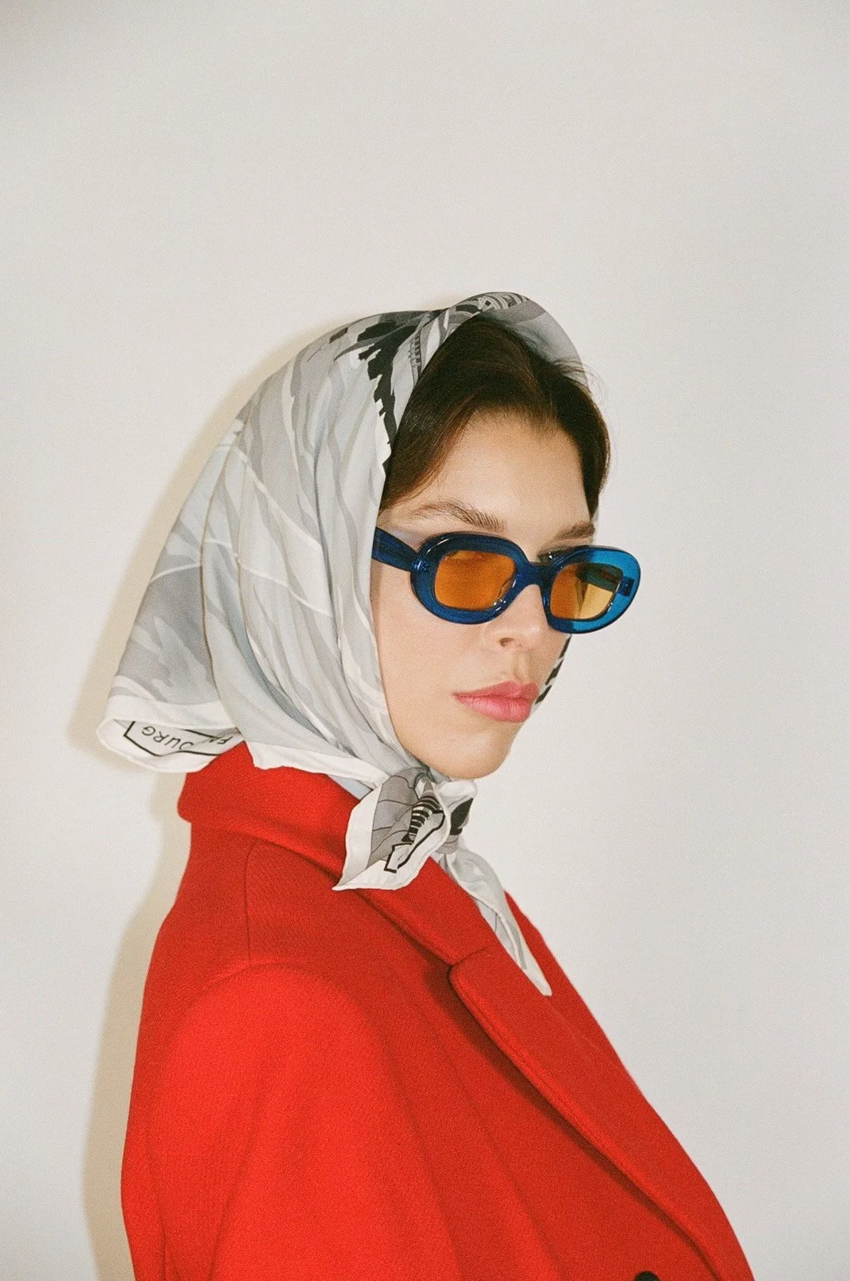 A woman with short dark hair wearing large blue sunglasses with orange lenses, a white patterned silk scarf on her head, and a red jacket, posed against a plain white background.