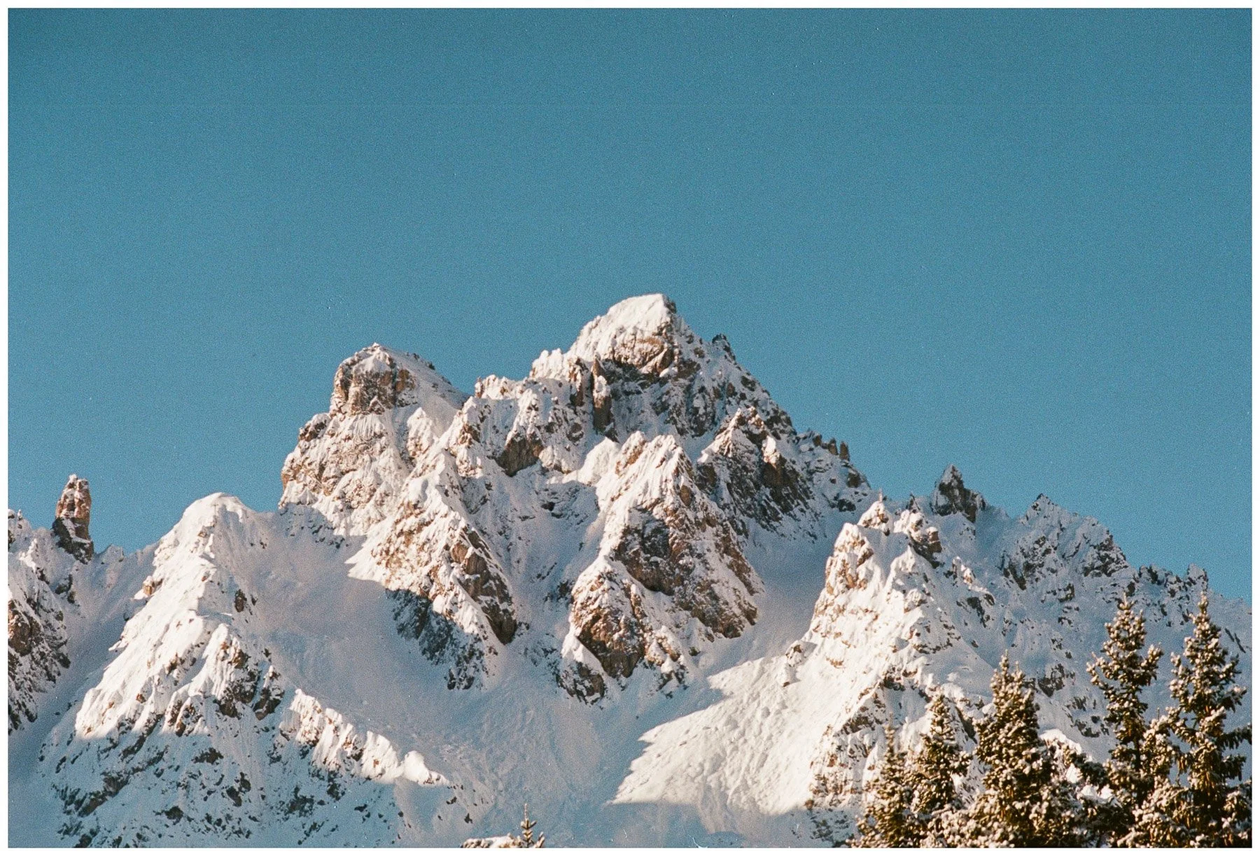 Snow-covered mountain peaks against a clear blue sky, with some evergreen trees in the foreground.