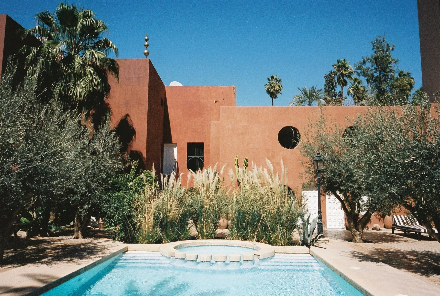 A swimming pool surrounded by greenery and trees, with a reddish-orange modern building in the background featuring round windows and a stone tower with decorative finials, under a clear blue sky.