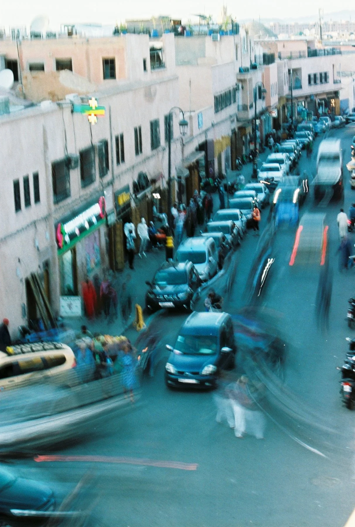 Street scene with parked cars and people walking along sidewalk in front of stores, blurry motion indicating movement.