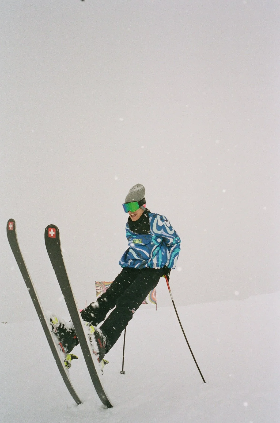 A person skiing in a snowy landscape, wearing a colorful jacket, black pants, a gray hat, and green ski goggles, appearing to be mid-air or performing a jump.