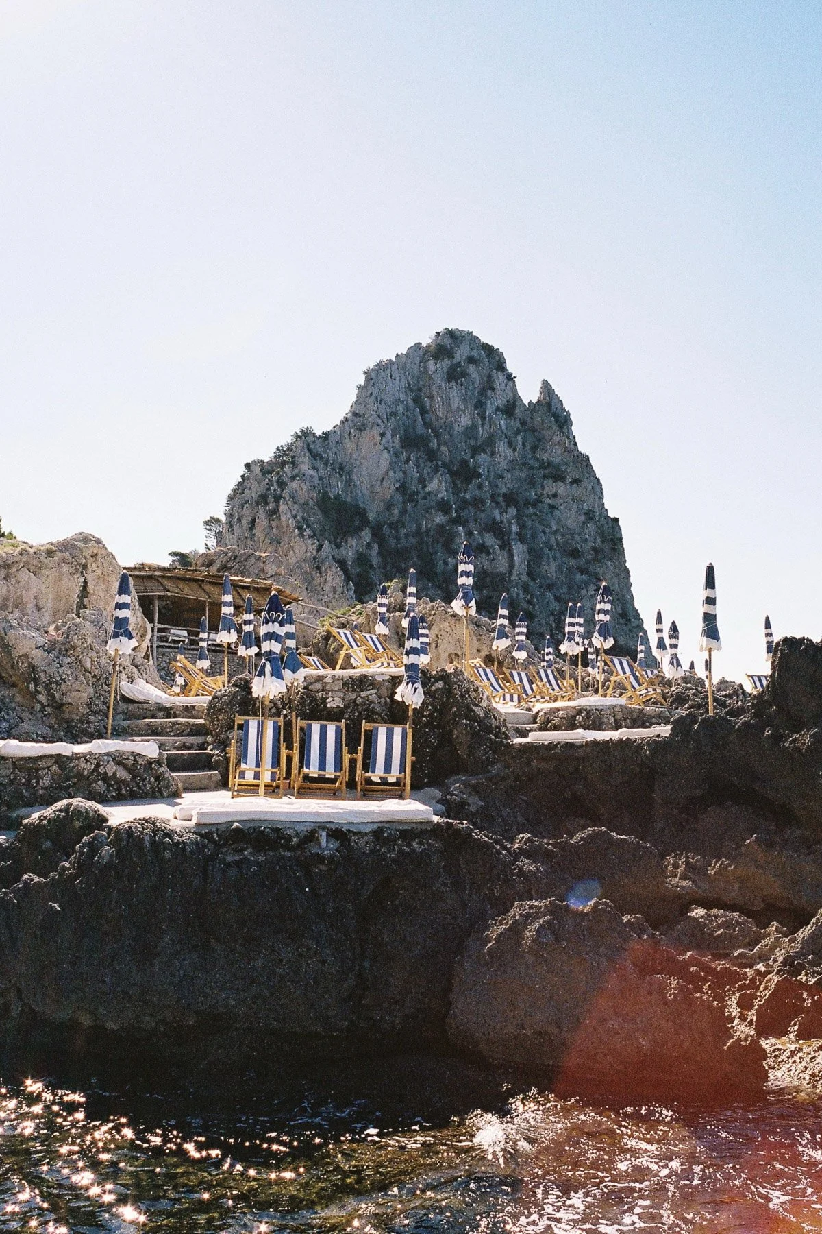 Beach with striped blue and white umbrellas and lounge chairs on rocky terrain, overlooking the sea with a large rocky mountain in the background under a clear sky.