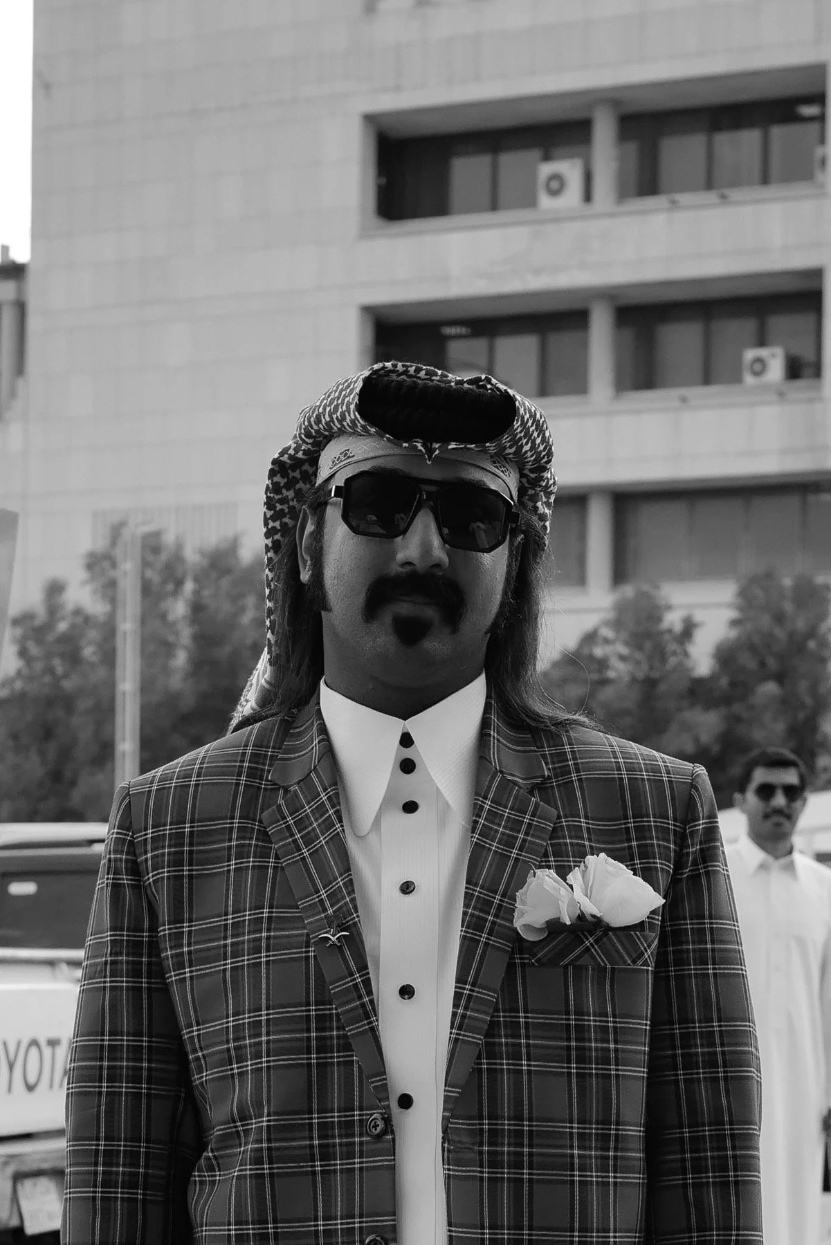 A man wearing a plaid suit jacket, white shirt with black buttons, a boutonniere, dark sunglasses, and a headscarf stands outdoors with a building and trees in the background.