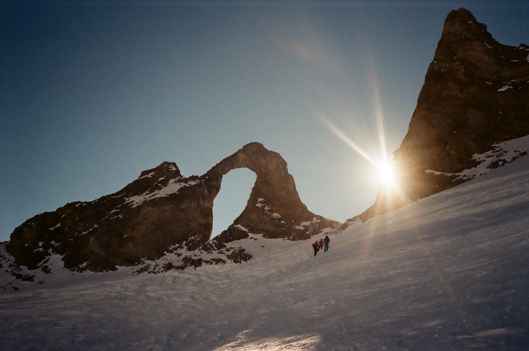 Snow-covered mountain landscape with a natural rock arch and sunburst behind the mountains. Small groups of hikers in colorful gear trekking through the snow.