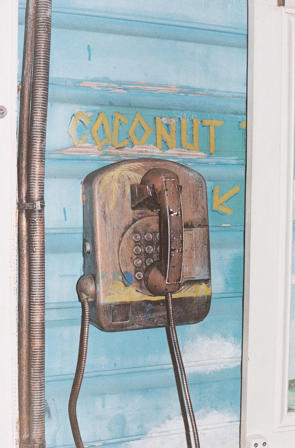 Old wooden payphone with a rotary dial, mounted on a colorful blue wall with the word 'COCONUT' painted in yellow above it. An arrow points toward the phone.