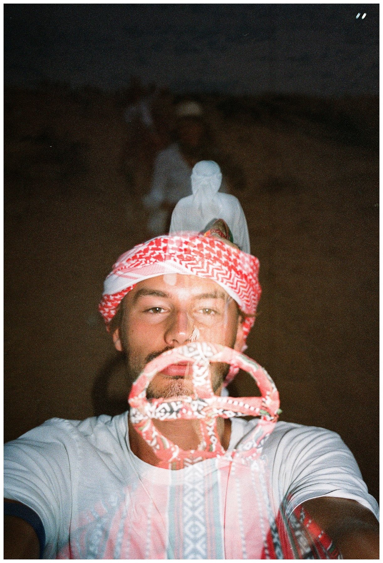 A man taking a selfie outdoors at night, wearing a traditional red and white keffiyeh and holding a patterned head covering over his face, with a dark, arid landscape and a white stone statue in the background.