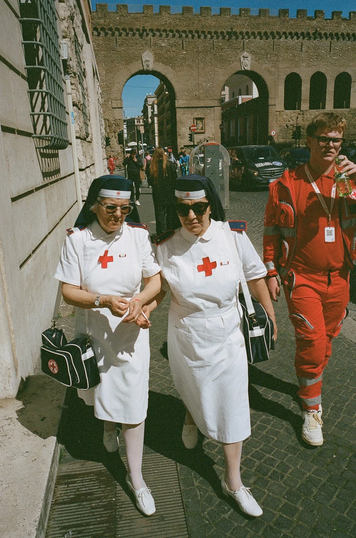 Three emergency medical personnel walking on a cobblestone street near an old gate. Two are women wearing white dresses and nurse hats with red crosses, carrying black medical bags. The third person is a man in a red medical uniform. The scene is in 