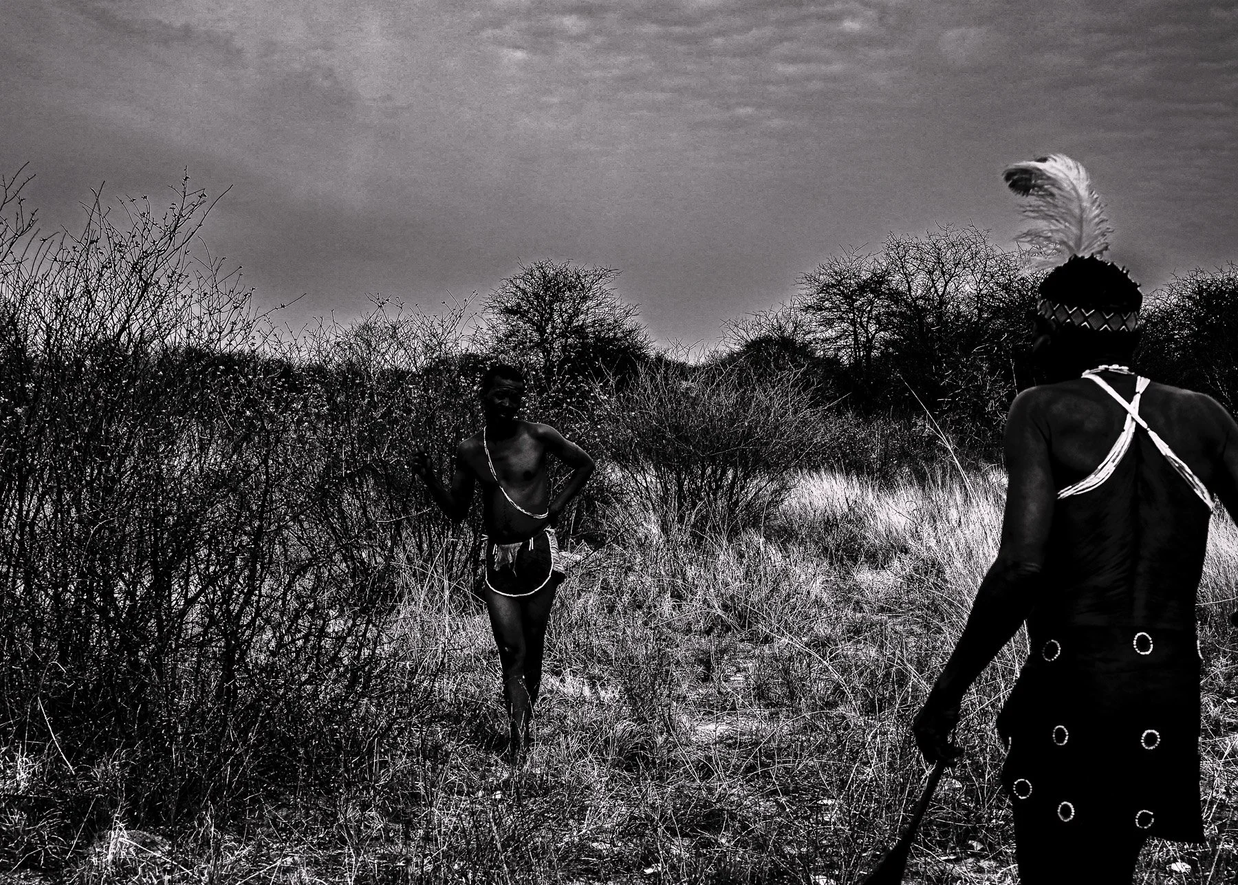 In a black-and-white photo, two indigenous men with traditional attire stand in a dry, bushy landscape with leafless trees. One man is seen from the back, wearing a skirt with circular patterns, and a feathered headpiece. The other man, shirtless wit