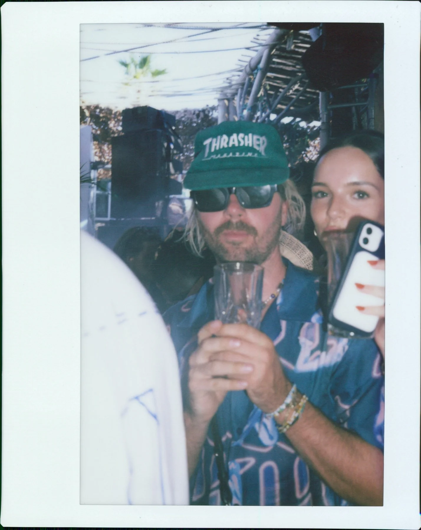 Two people taking a selfie at a party or gathering. The man in the center wears a green Thrasher hat, sunglasses, and holds a drink. The woman on the right holds a phone with a white and orange case, smiling at the camera.