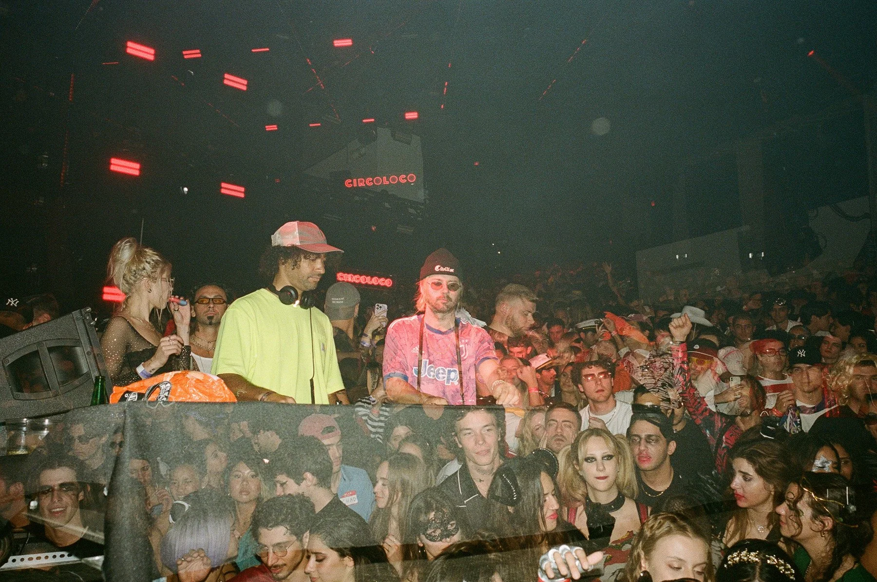 Crowd of people at a nightclub or concert with a DJ at the front, neon signs that read 'Circoloco,' and colorful lighting.