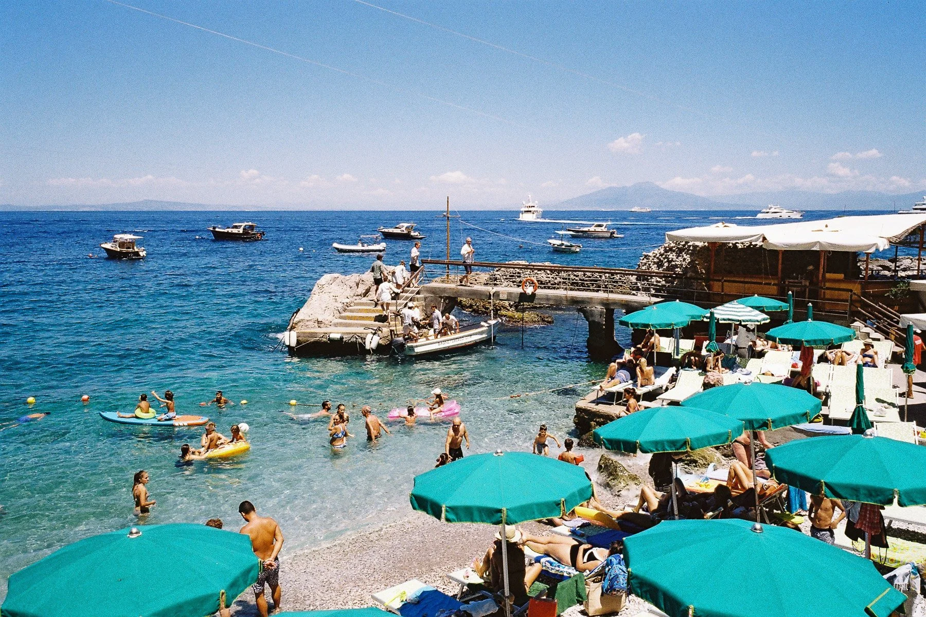 Beach scene with people swimming and lounging under green umbrellas, boats on the water, and a pier with more people and boats.