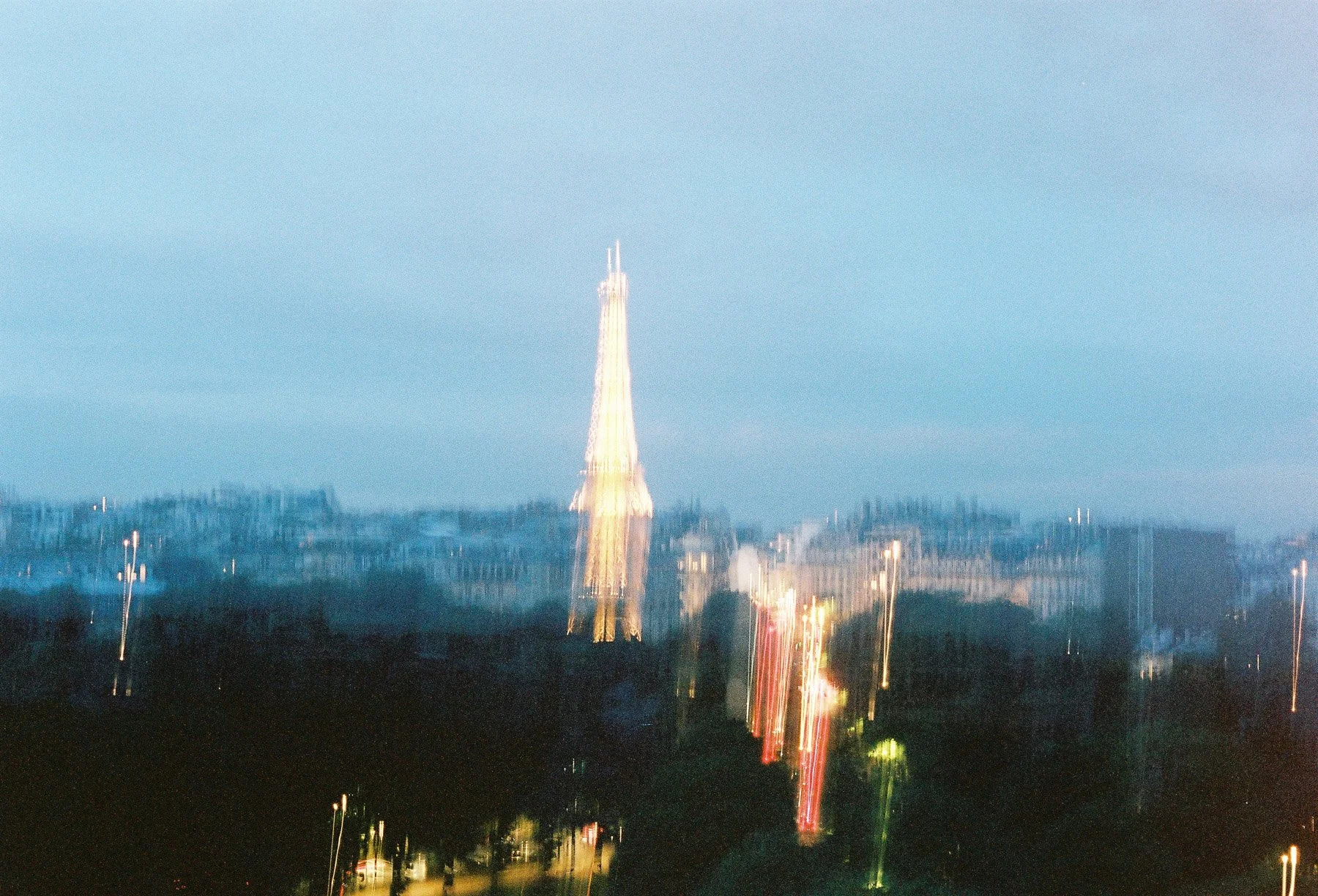 Nighttime cityscape featuring a tall illuminated tower, possibly the Eiffel Tower, with colorful light reflections and blurred city lights in the background.