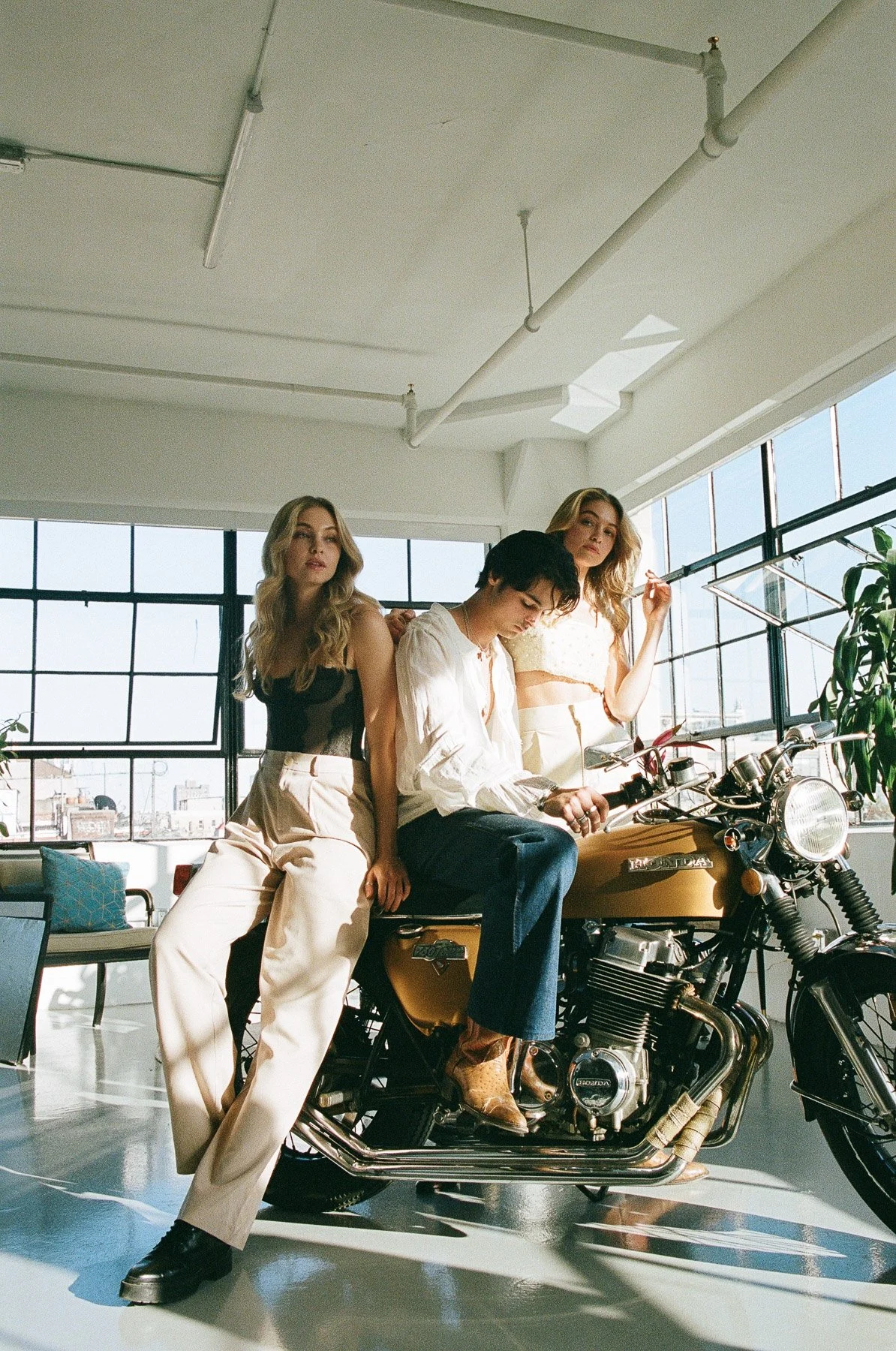 Three young women with light skin posing in an industrial-style studio with large windows and a motorcycle. One woman is sitting on the motorcycle, and the other two are standing beside her.