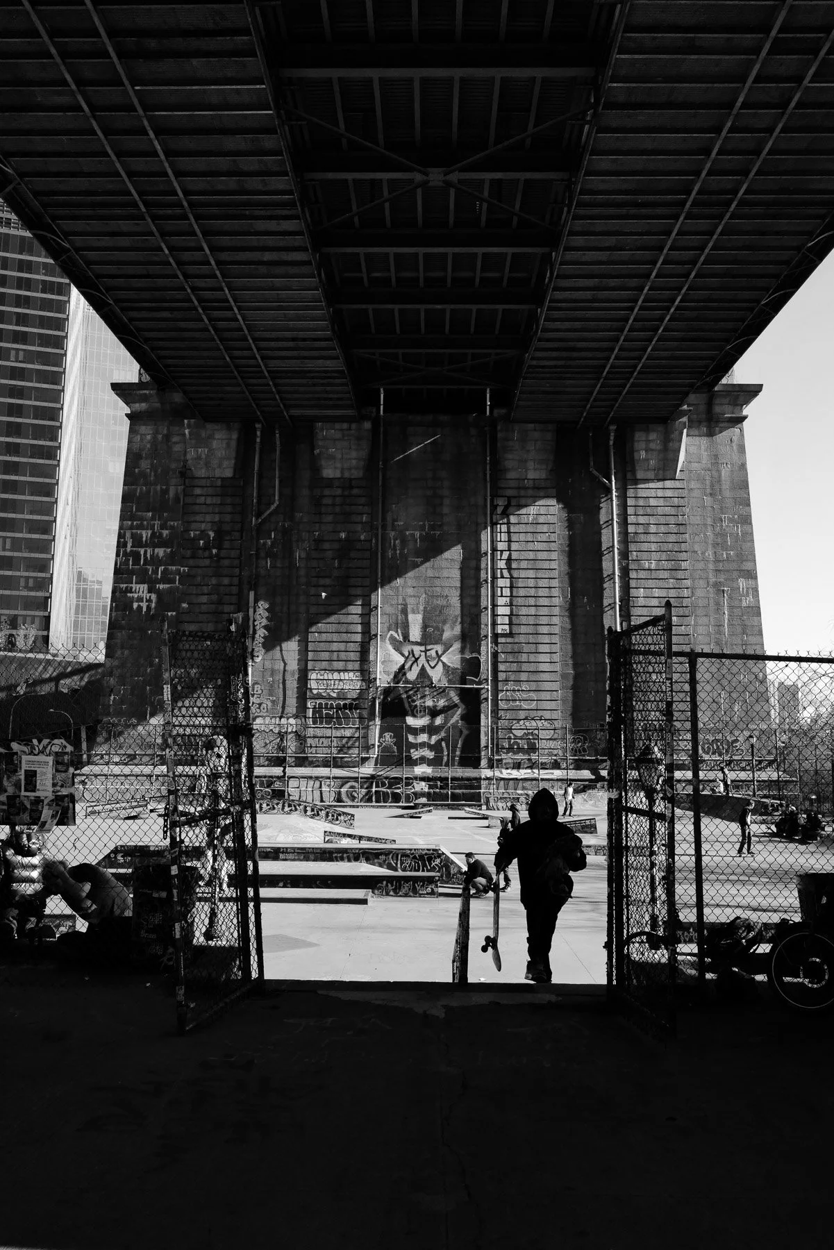 Under a bridge in an urban park, a fenced skatepark is visible with graffiti on the walls. Several people are gathered, some sitting, some standing, and one person walking out of the gate. Skateboards are on the ground.