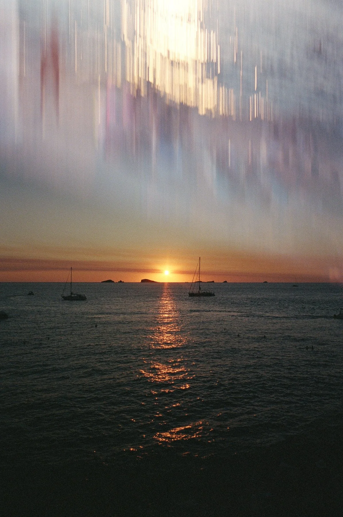A sunset over the ocean with sailboats and distant islands, with the sky reflecting on the water and a dramatic cloud formation above.
