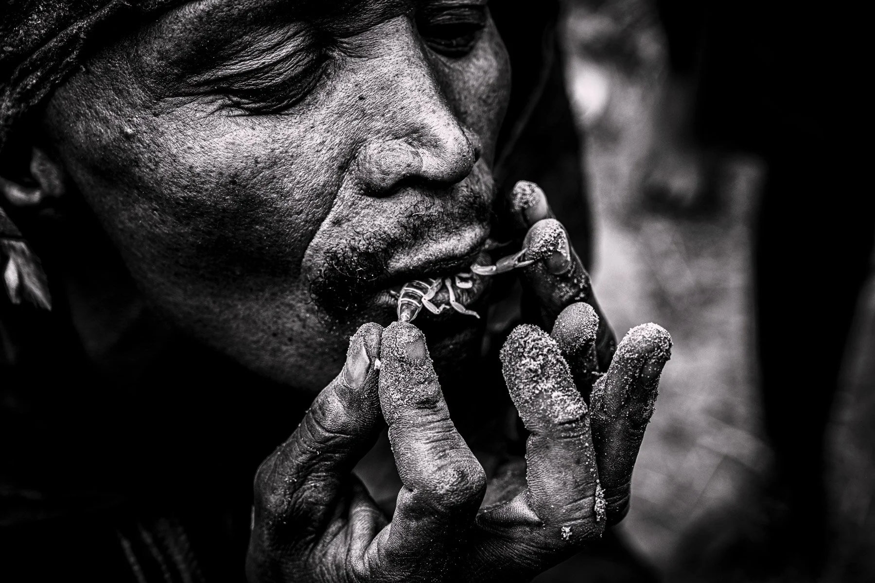 Close-up of an elderly person with dark skin, tightly holding a small figurine or object with their fingers covered in a powdery substance. The person's face is partially visible, with a focus on their closed eyes and textured skin.