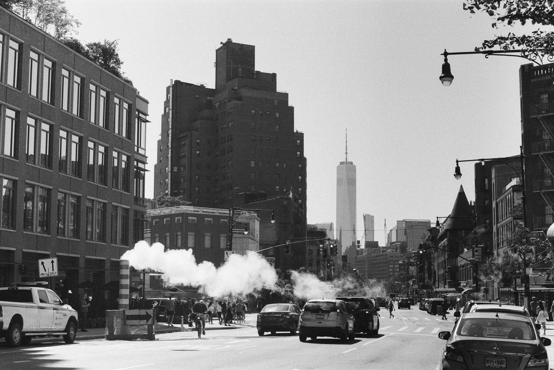 Street scene in New York City with buildings, cars, pedestrians, steam rising from underground, and One World Trade Center in the background.