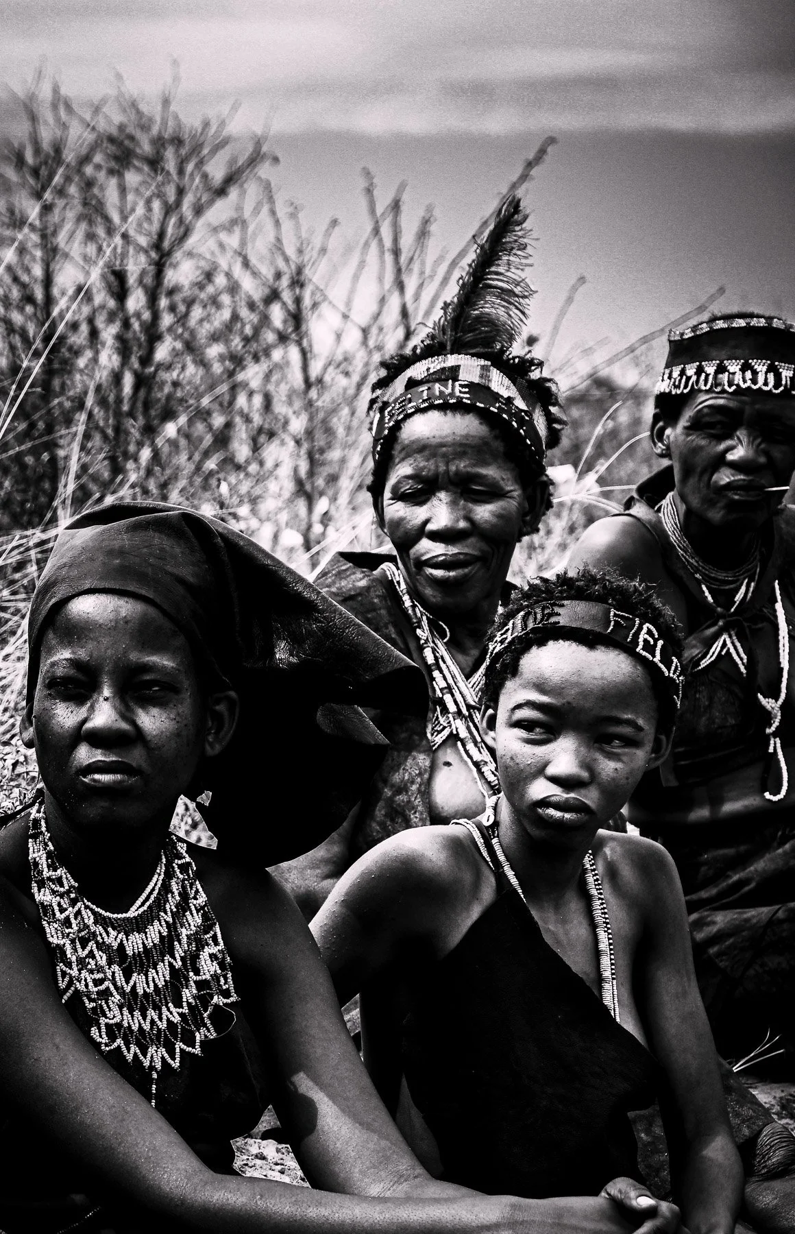 Black and white photograph of four indigenous African women and girls wearing traditional beaded jewelry and headbands, sitting outdoors with a background of dry grass and trees.