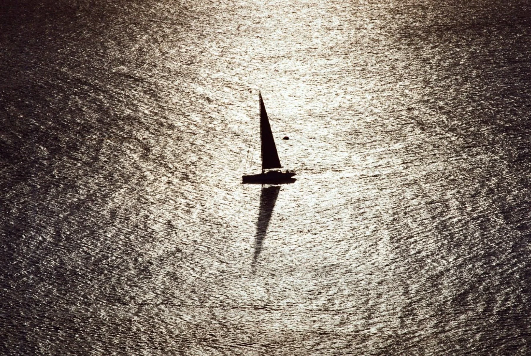 Silhouette of a sailboat on a shimmering water surface with the sun reflecting off the water, creating a sparkling backdrop.
