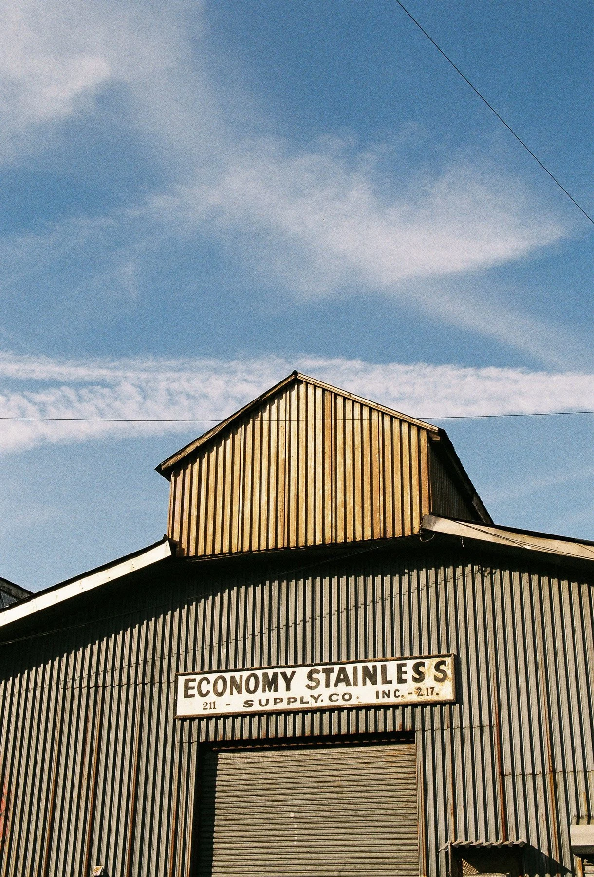 A rustic building with a corrugated metal facade and a wooden section on top, featuring a sign that reads 'Economy Stainless Supply Co., Inc.' The sky is blue with scattered clouds, and power lines are visible overhead.