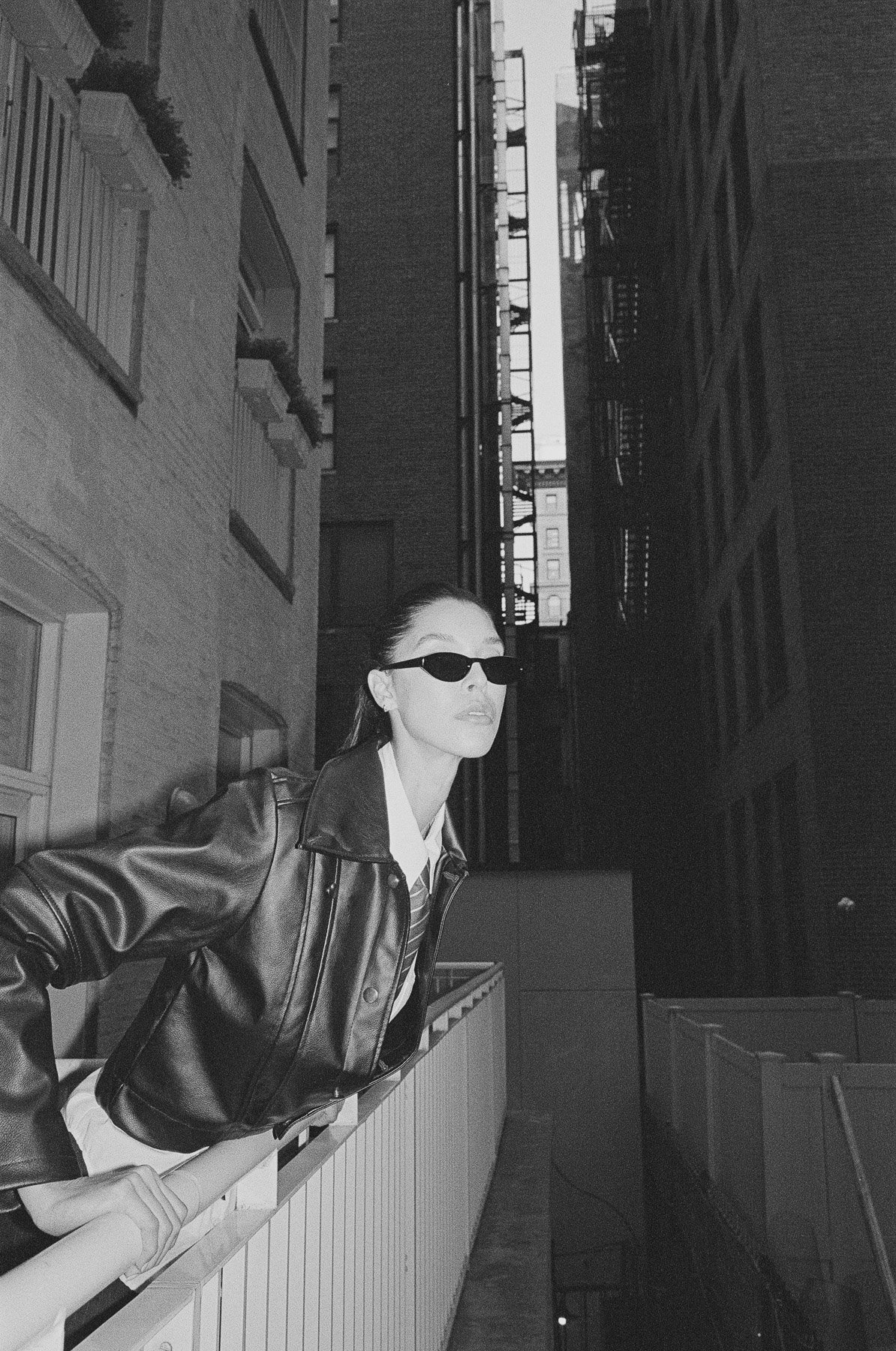 A woman wearing sunglasses and a leather jacket leaning on a balcony railing in an urban setting during daytime.