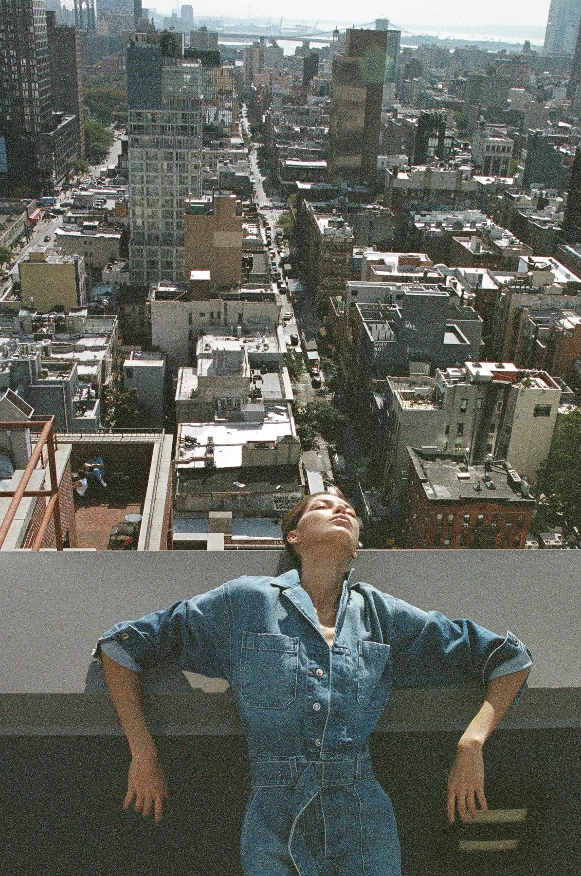 A woman in a denim jumpsuit leaning back with her eyes closed and arms outstretched on a rooftop, overlooking a cityscape with tall buildings and distant water or sky in the background.