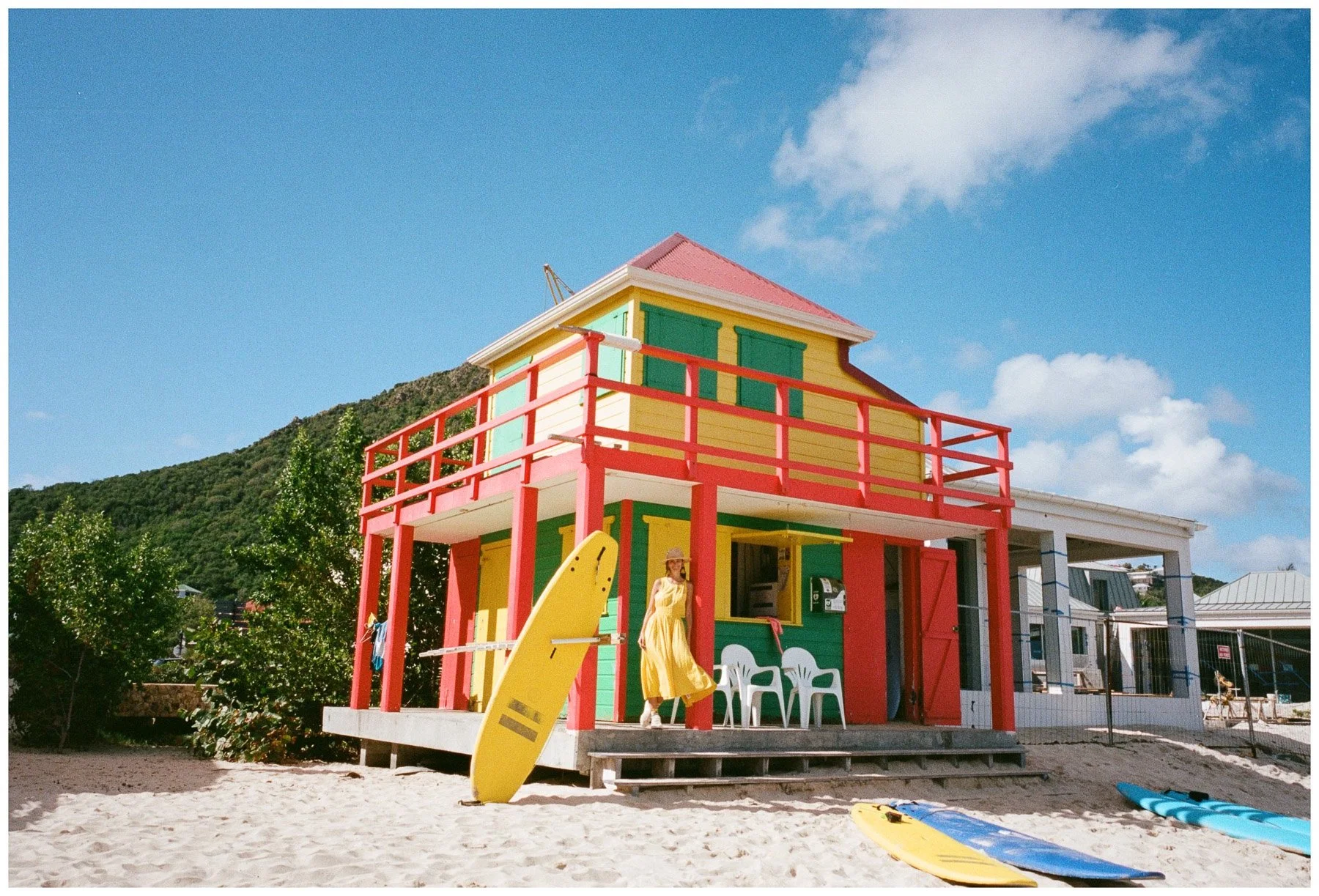 Colorful beach house with a yellow exterior, red balcony, and green window shutters, located on a sandy beach. There are surfboards on the sand, and a woman in a yellow dress and sun hat walking nearby. The sky is blue with a few clouds, and some gre