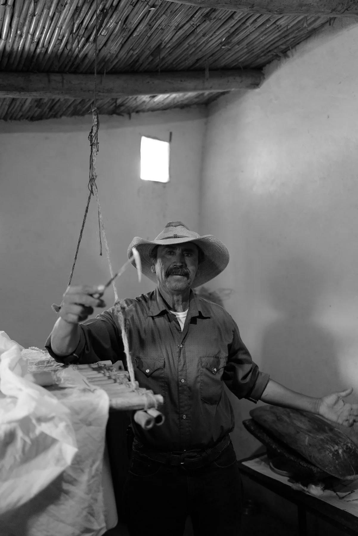 A man with a mustache is wearing a wide-brimmed hat and a button-up shirt, standing indoors with his arms outstretched. The room has awooden or bamboo ceiling with a small window in the background. The man appears to be demonstrating or explaining so