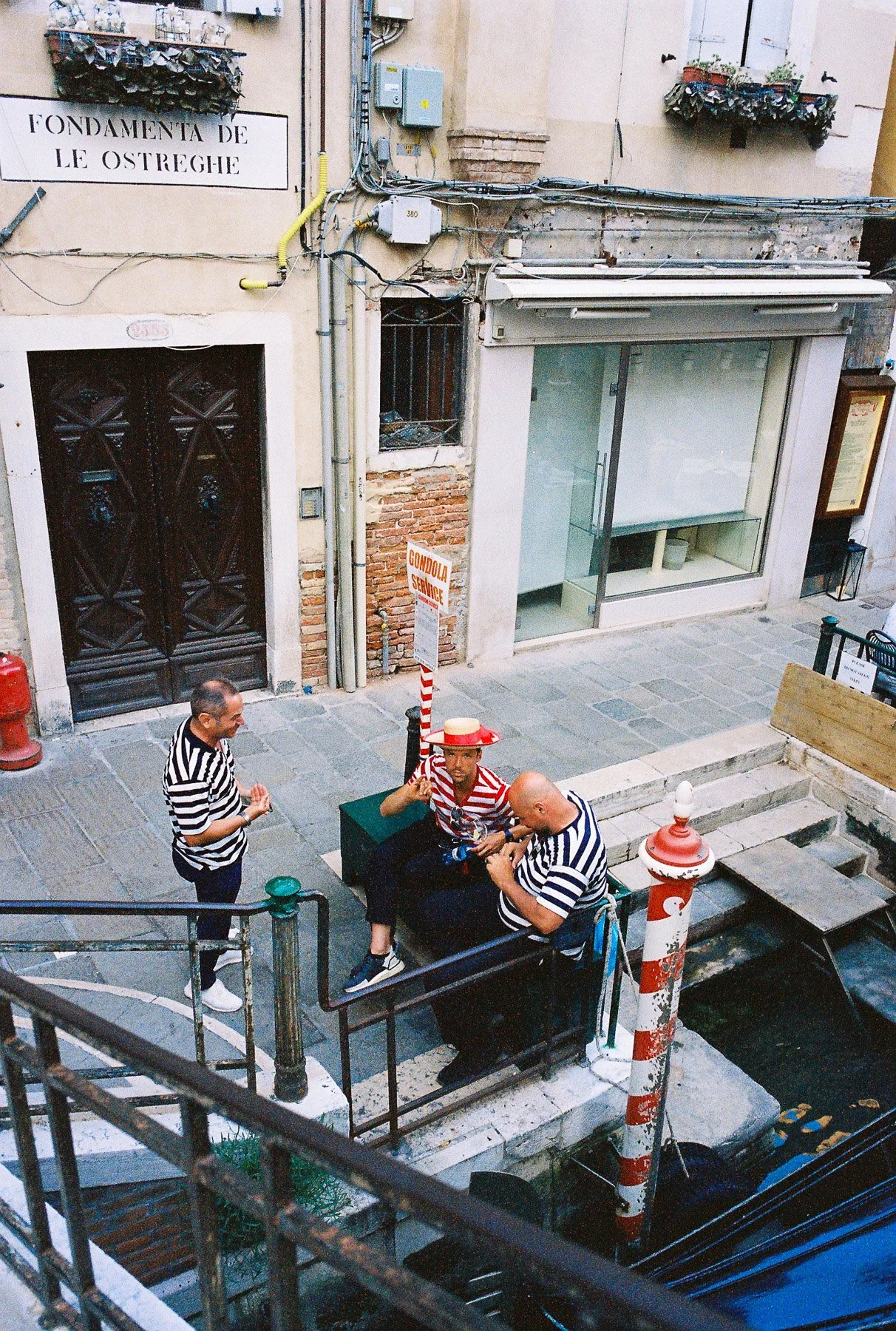 Three men in striped shirts are gathered on a small outdoor staircase, with one of them dressed in a red and white striped hat. They are near a canal with boats and a sign reading 'Gondola Service' in Italian. The surroundings have old buildings with
