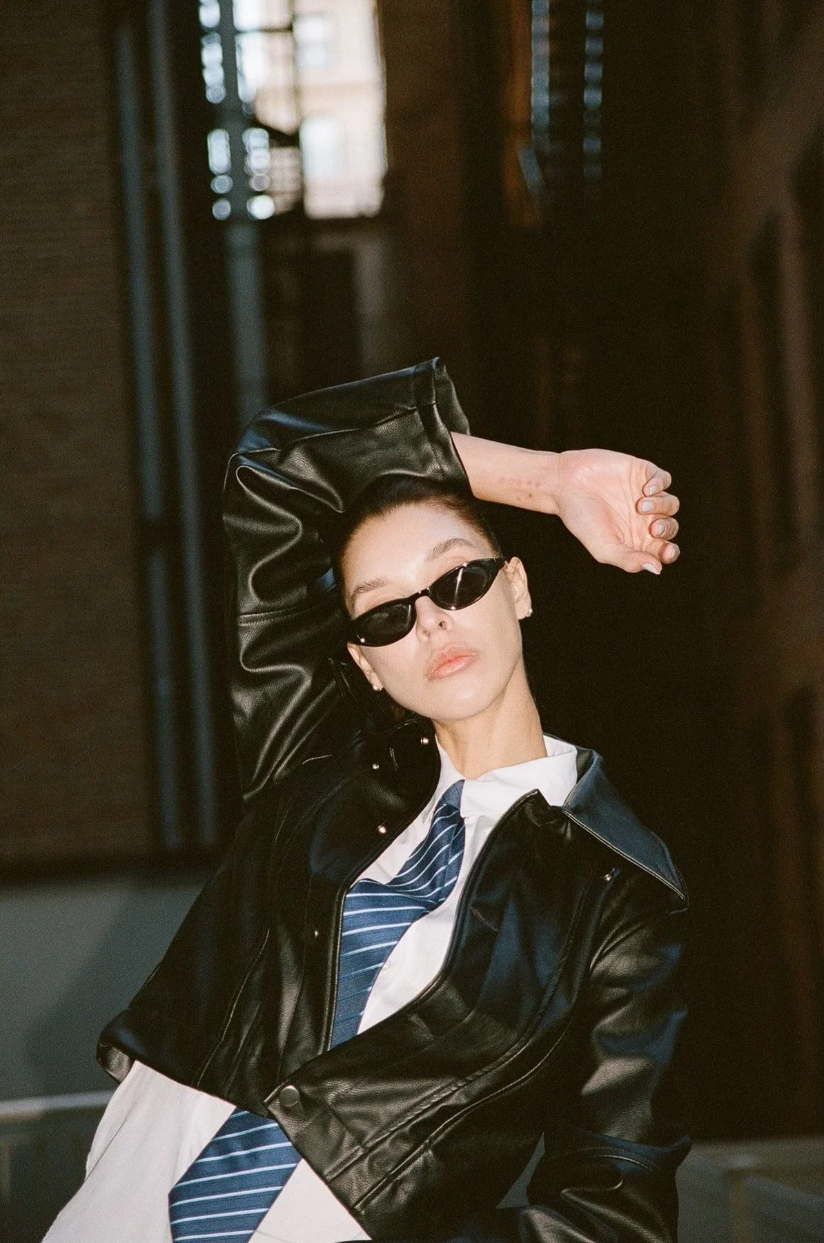 A woman wearing a leather jacket, striped tie, white shirt, and small black sunglasses, posing with her arm raised behind her head in front of a mirror.