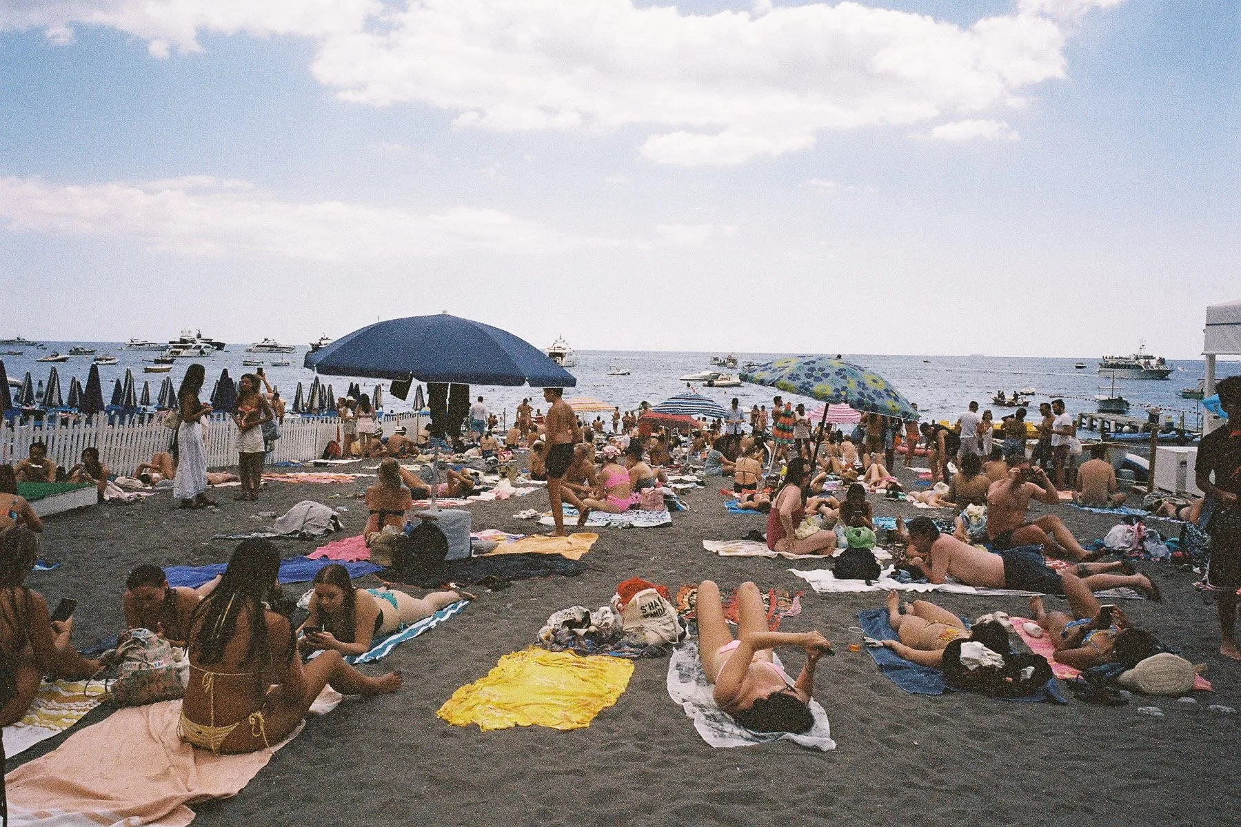 Crowded beach scene with many people sunbathing, sitting, and standing on the sand. Bright umbrellas are scattered across the beach, and boats are visible on the water. The sky is partly cloudy.