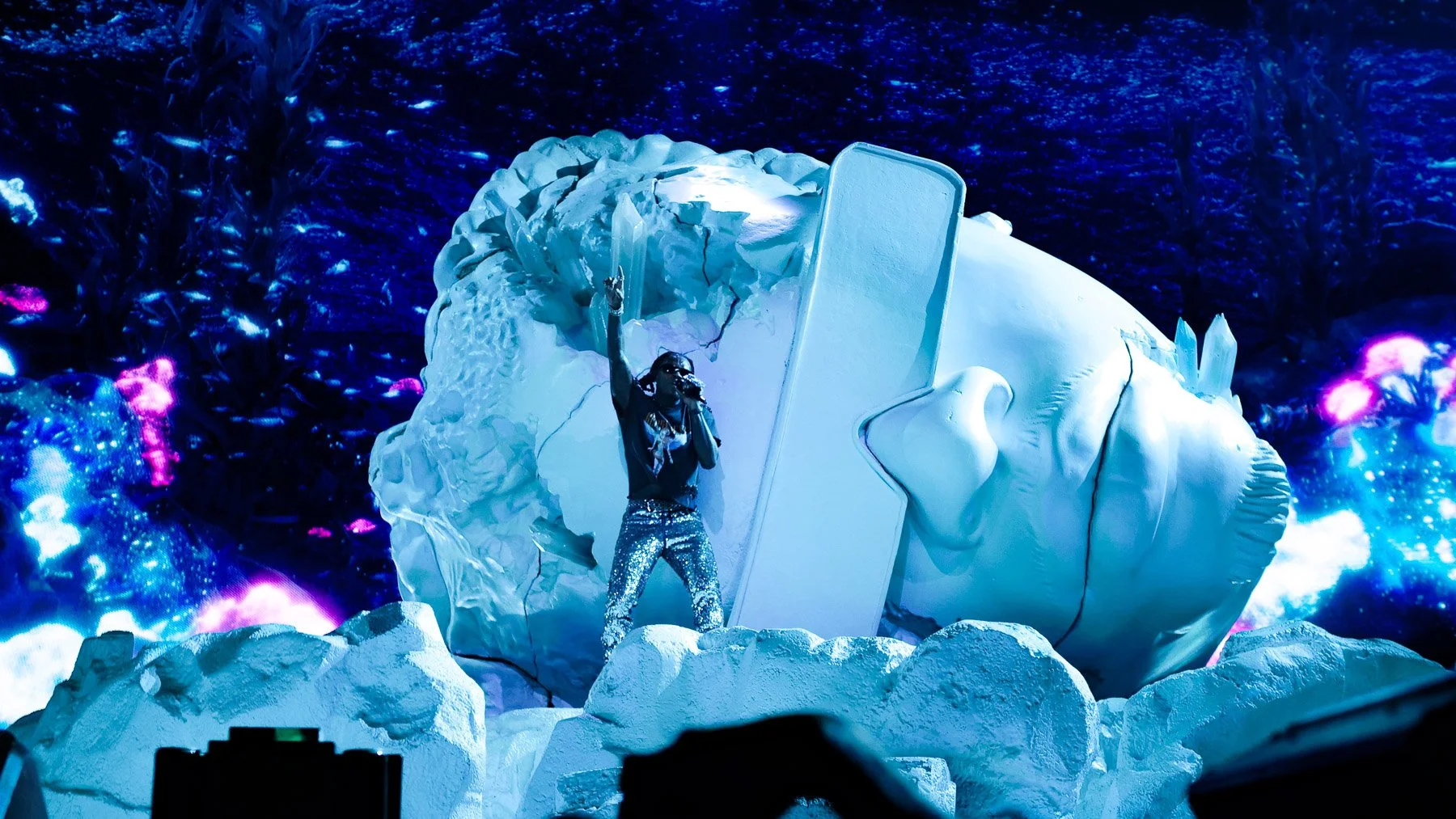 A performer wearing a black top and shiny pants holds a microphone and raises one arm in front of a large, white iceberg sculpture. The background features blue and purple lighting effects resembling undersea scenery.