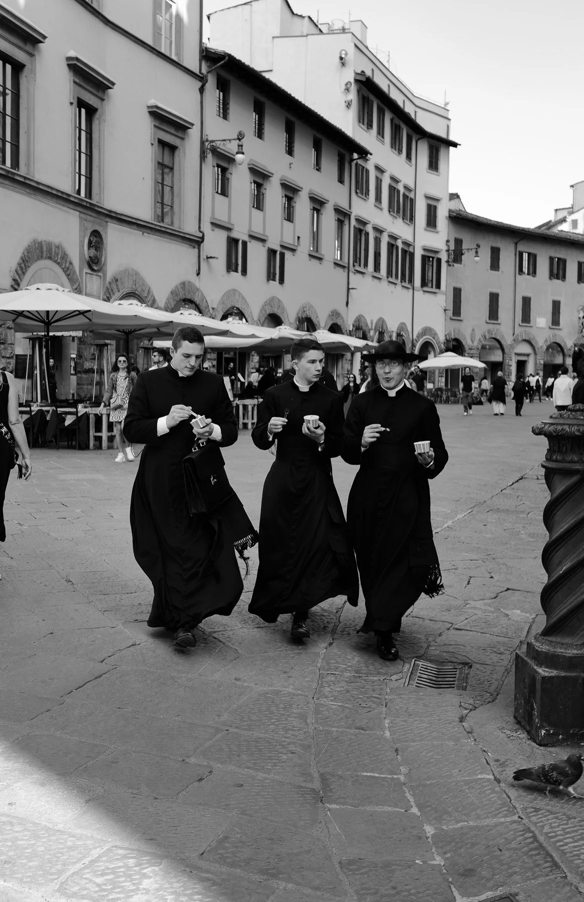 Three Catholic priests in black cassocks walking in a town square, holding cups of ice cream, with buildings and outdoor tables in the background.
