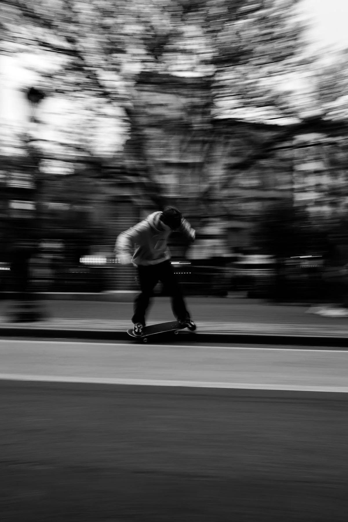 A person skateboarding on a street in black and white. The background shows trees and a building with a motion blur effect.