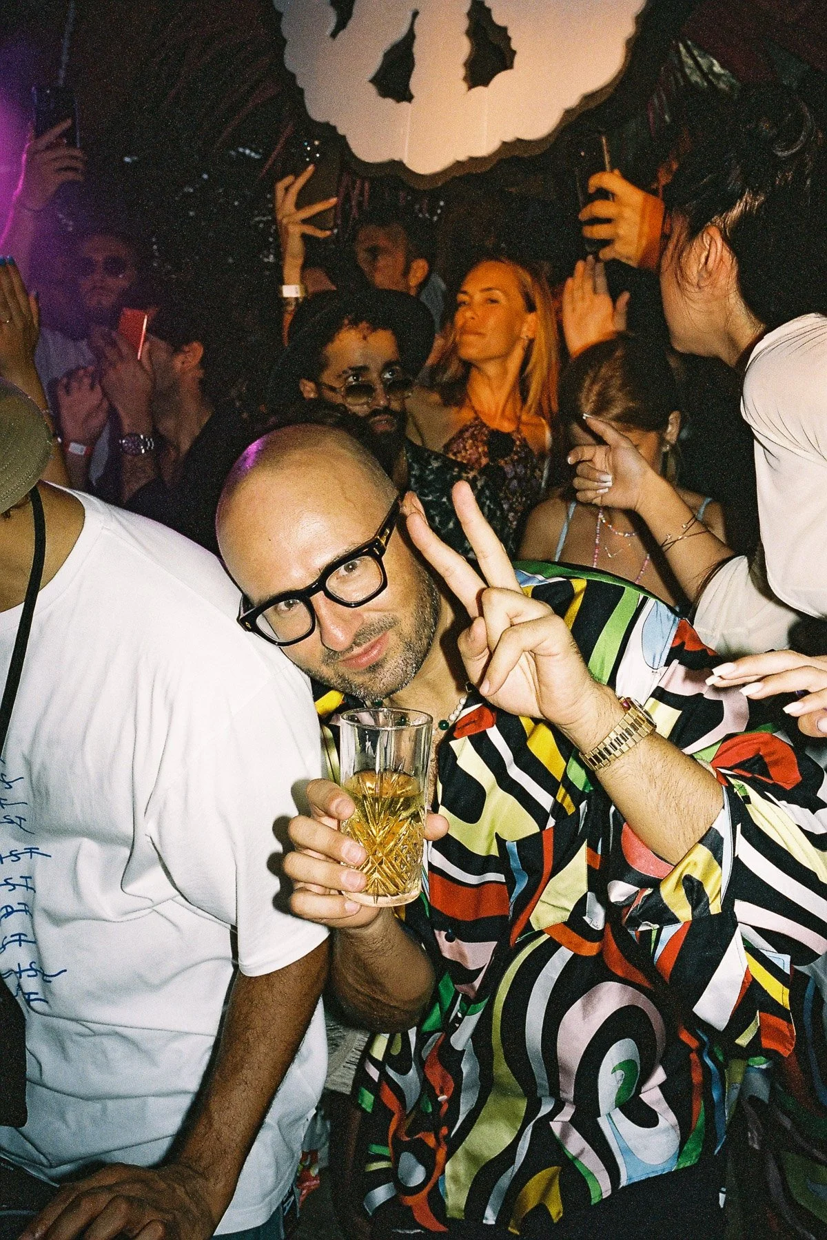 A man with glasses holding a drink and making a peace sign, surrounded by people at a party or celebration with colorful lighting and decorations.
