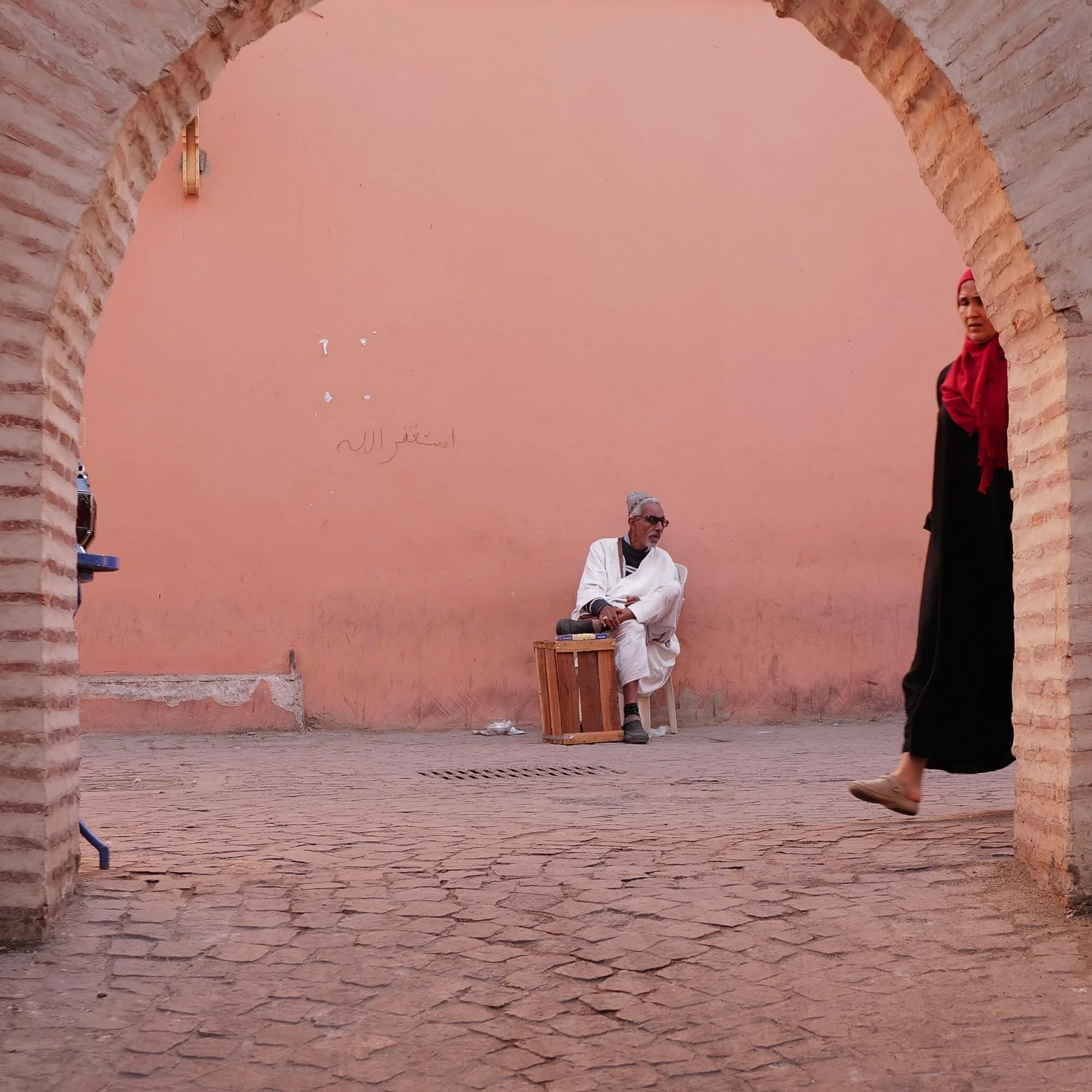 A man with gray hair and glasses sitting on a white plastic chair, wearing traditional white attire, in a pink-walled courtyard with an archway. A woman wearing a red headscarf, black dress, and beige shoes is walking past the arch on the right. Ther