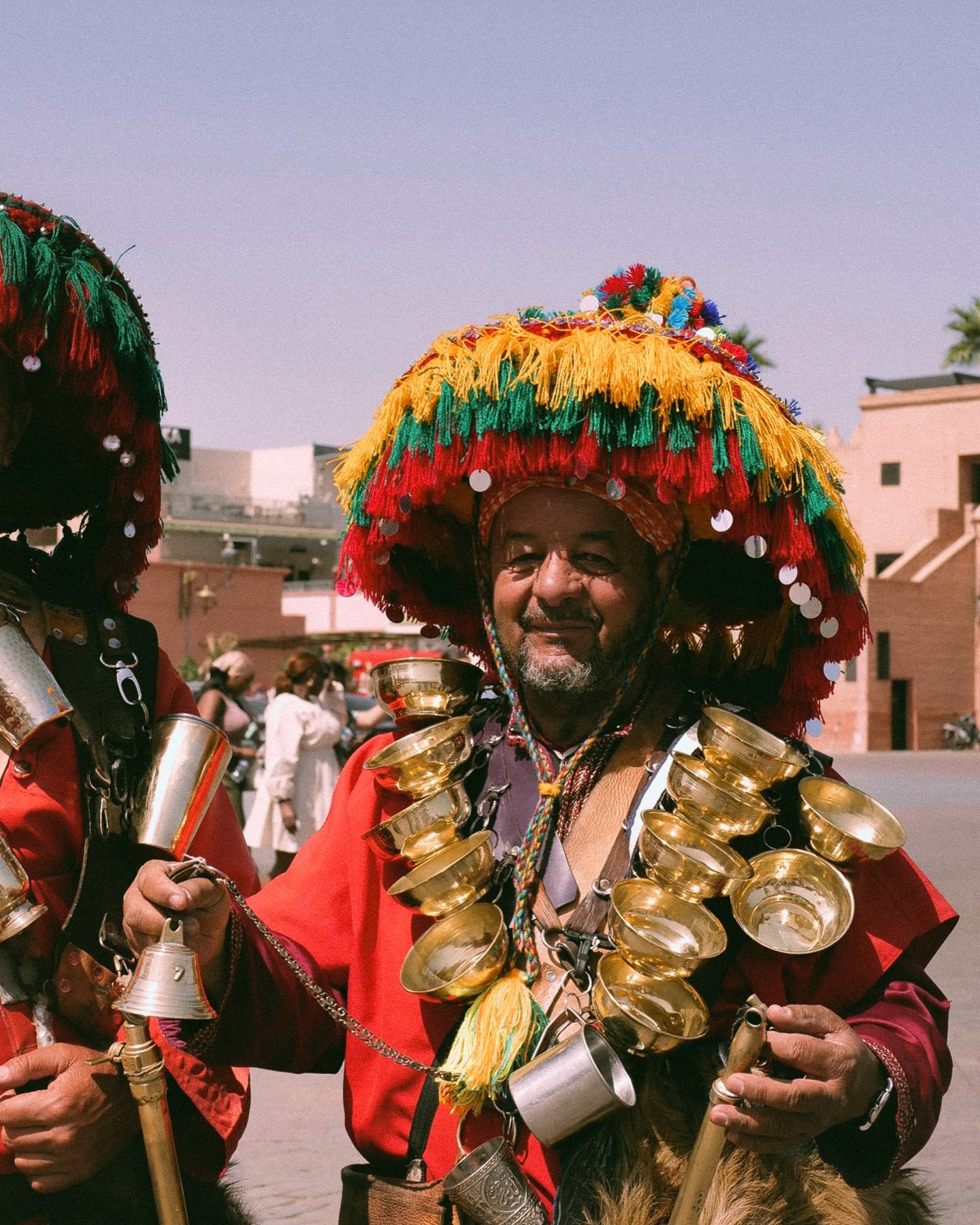 A man celebrating a festival wearing a colorful, fringed hat decorated with pom-poms and sequins. He is smiling and holding musical instruments around his neck and waist, dressed in traditional attire with bright red and other vibrant colors.