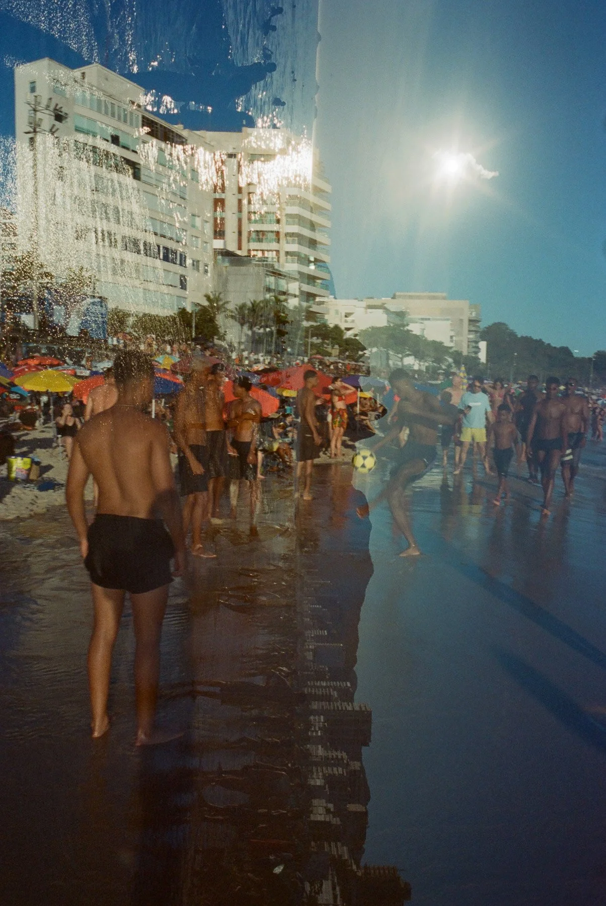 People swimming and relaxing on a crowded beach with umbrellas, high-rise buildings, and a clear sky with the sun shining.