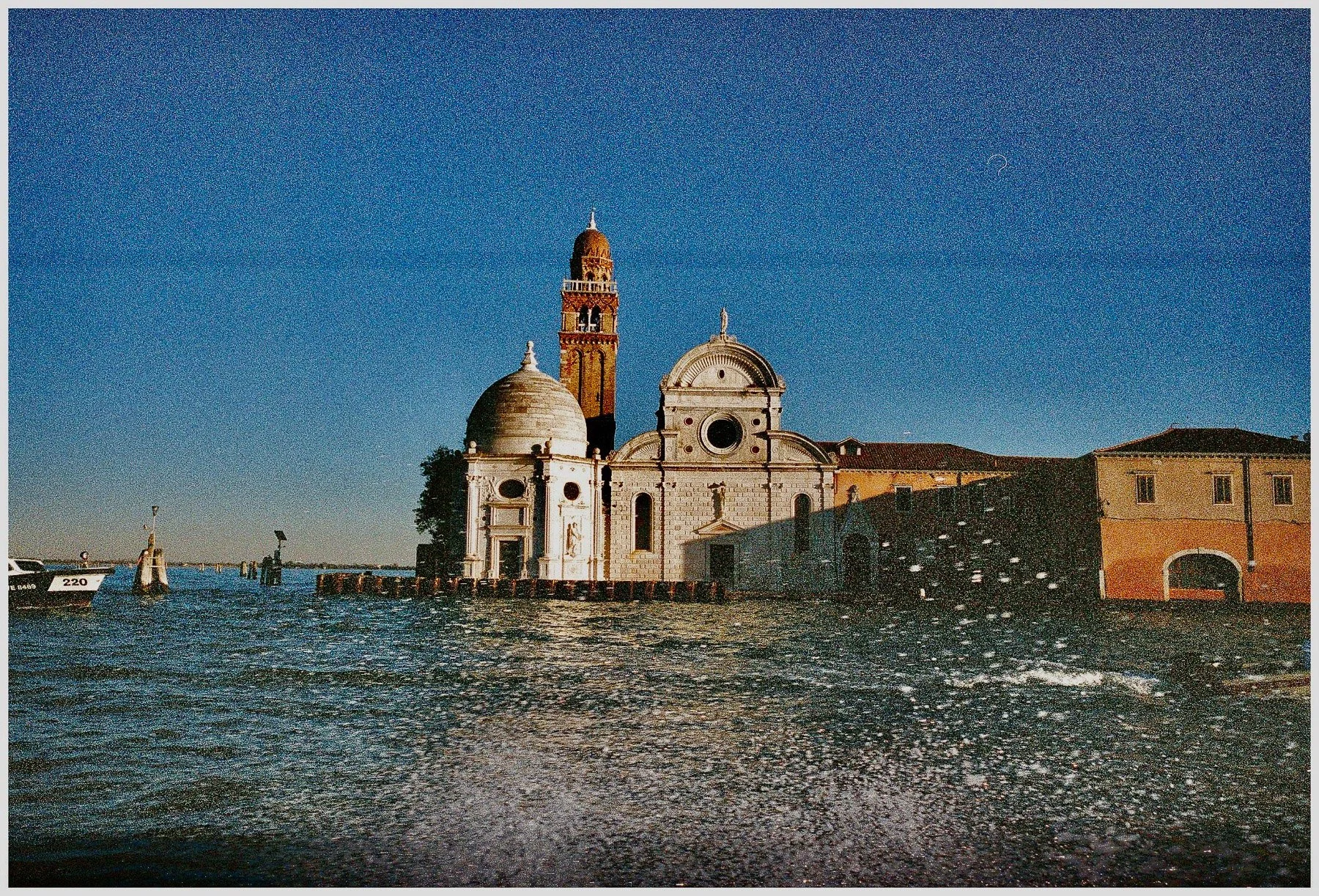 View of historic buildings along a waterfront at sunset, with a church and a bell tower visible, and boats docked on the water.
